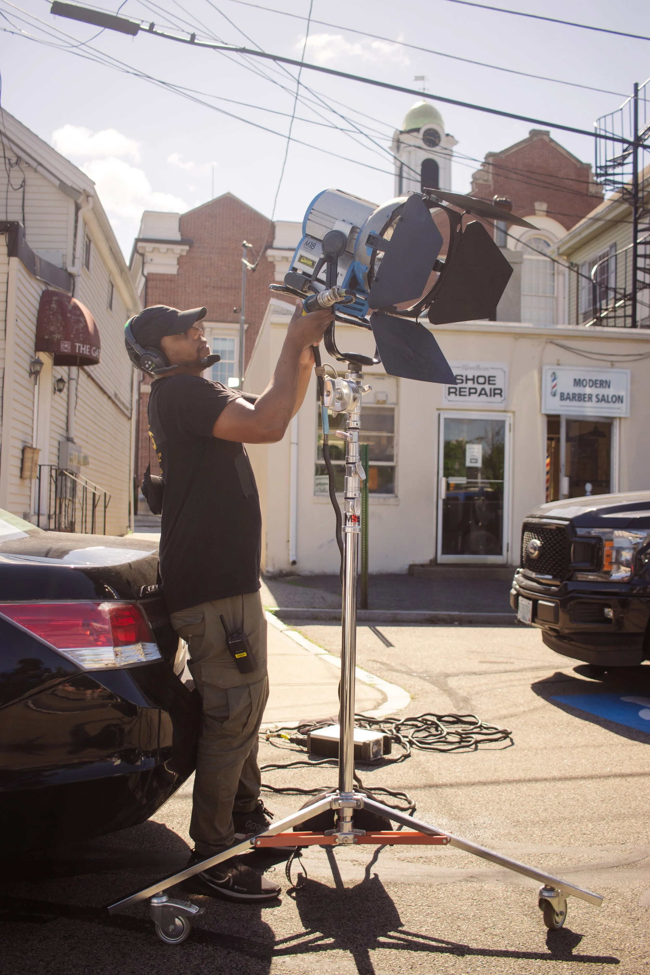 A man wearing a black cap and headset is operating a professional film or photography lighting equipment on a city street, with buildings and a church tower in the background.