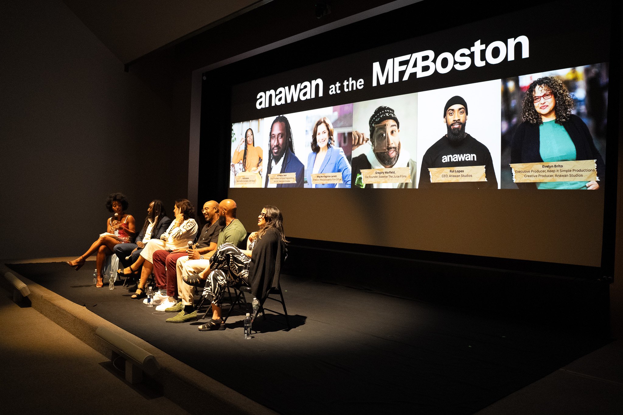 Panel of six diverse people sitting on chairs on a stage during a panel discussion, with a large screen behind them displaying headshots and names of panelists at the MFABoston event.