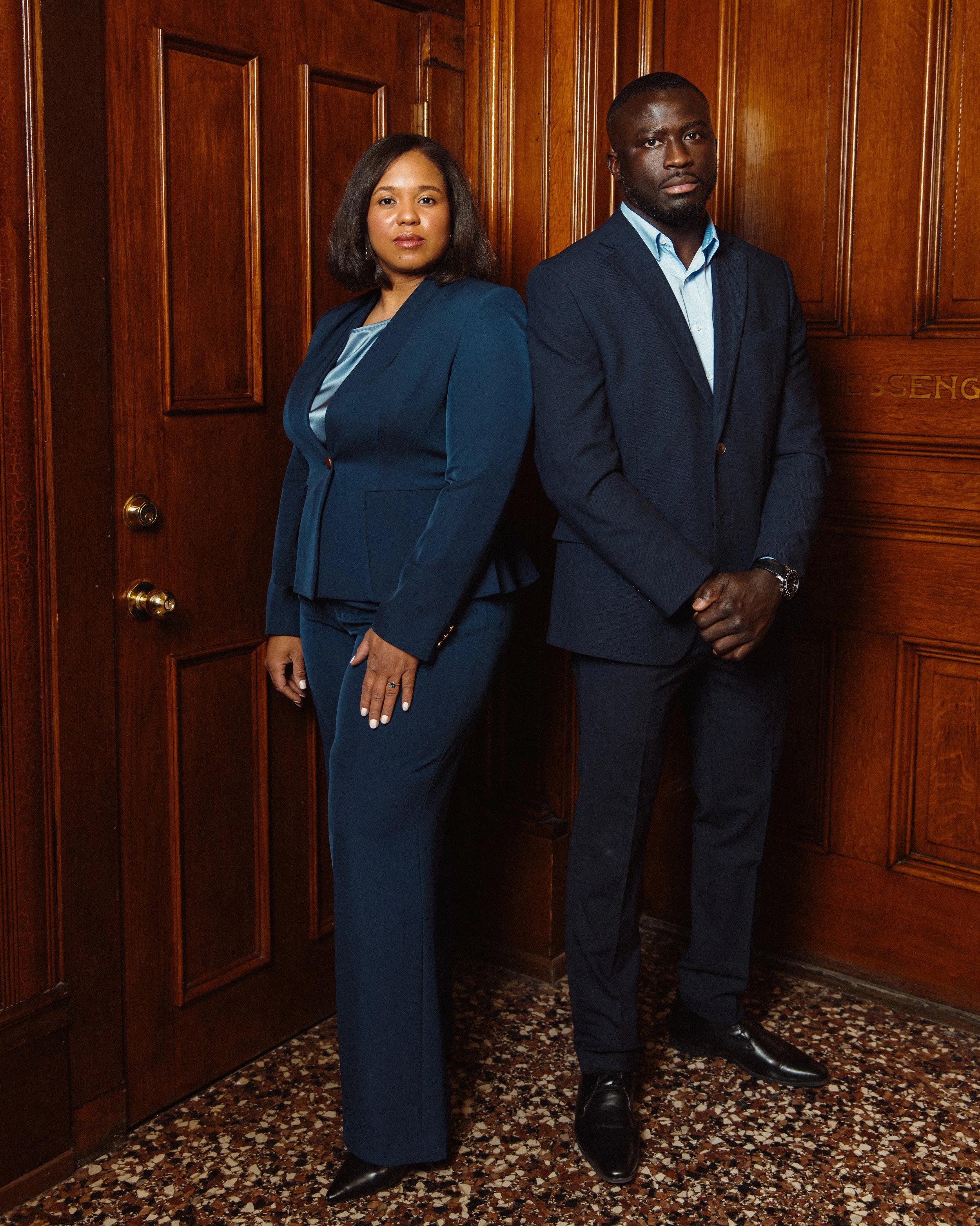 stand side by side indoors against a wooden paneled background. Both are dressed in dark blue suits, with the woman wearing a blazer and trousers, and the man in a blazer, shirt, and trousers. They have serious expressions, with the woman having her 
