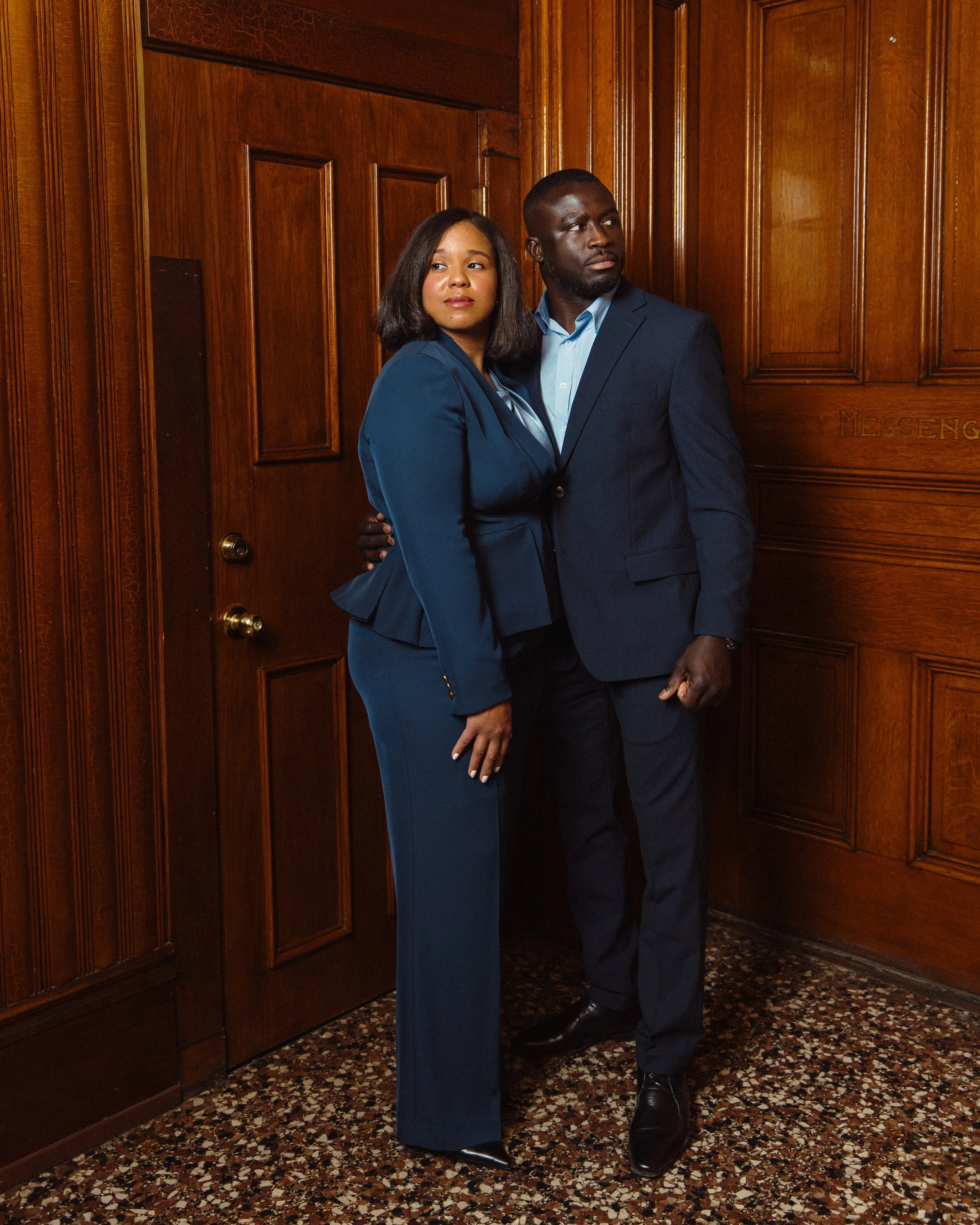 Francois & Iraimi Diatta of Empower Nutrition dressed in formal attire standing against a wooden paneled wall in an indoor setting with a terrazzo floor.