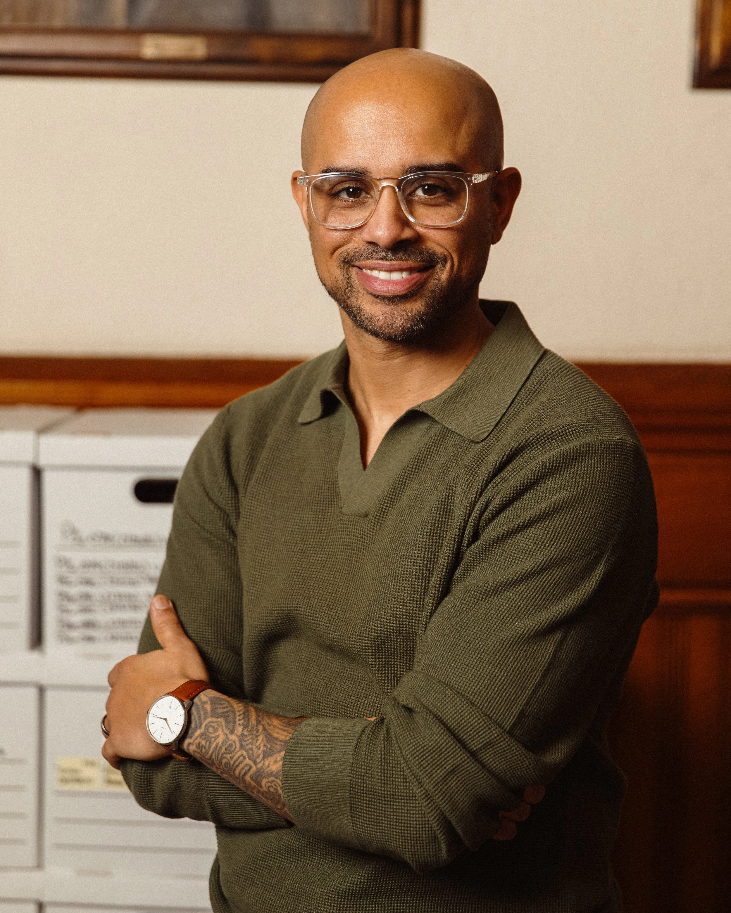 Nelson Fernandes, Deputy Chief of Staff with a beard and bald head, wearing glasses and a green sweater, standing with arms crossed, in a room with framed art on the wall and white furniture in the background.