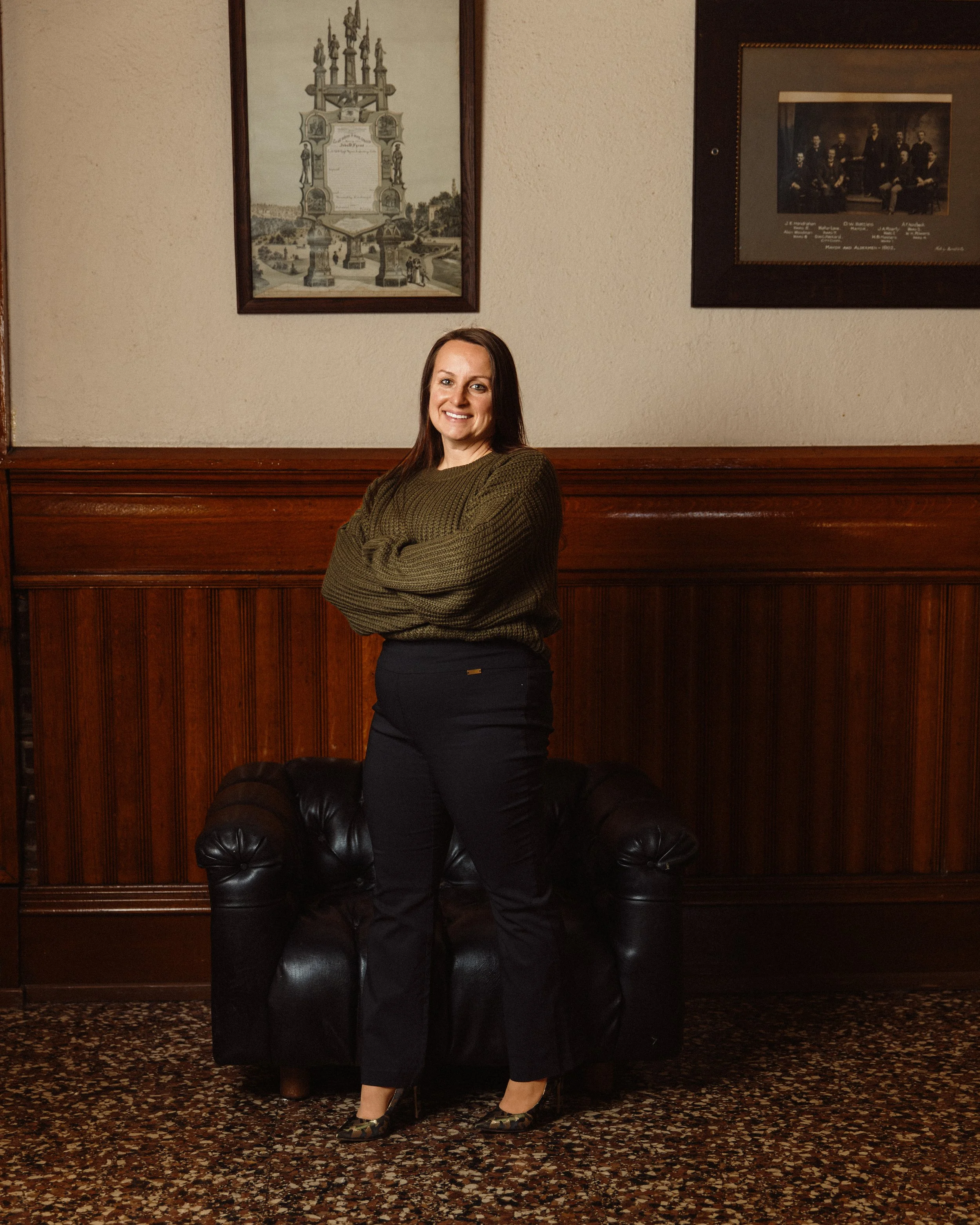 A woman standing in a room with wood-paneled walls, smiling and crossing her arms. She is wearing a green knit sweater, black pants, and patterned high heels. Behind her, there is a black leather couch, two framed pictures on the wall, and patterned 