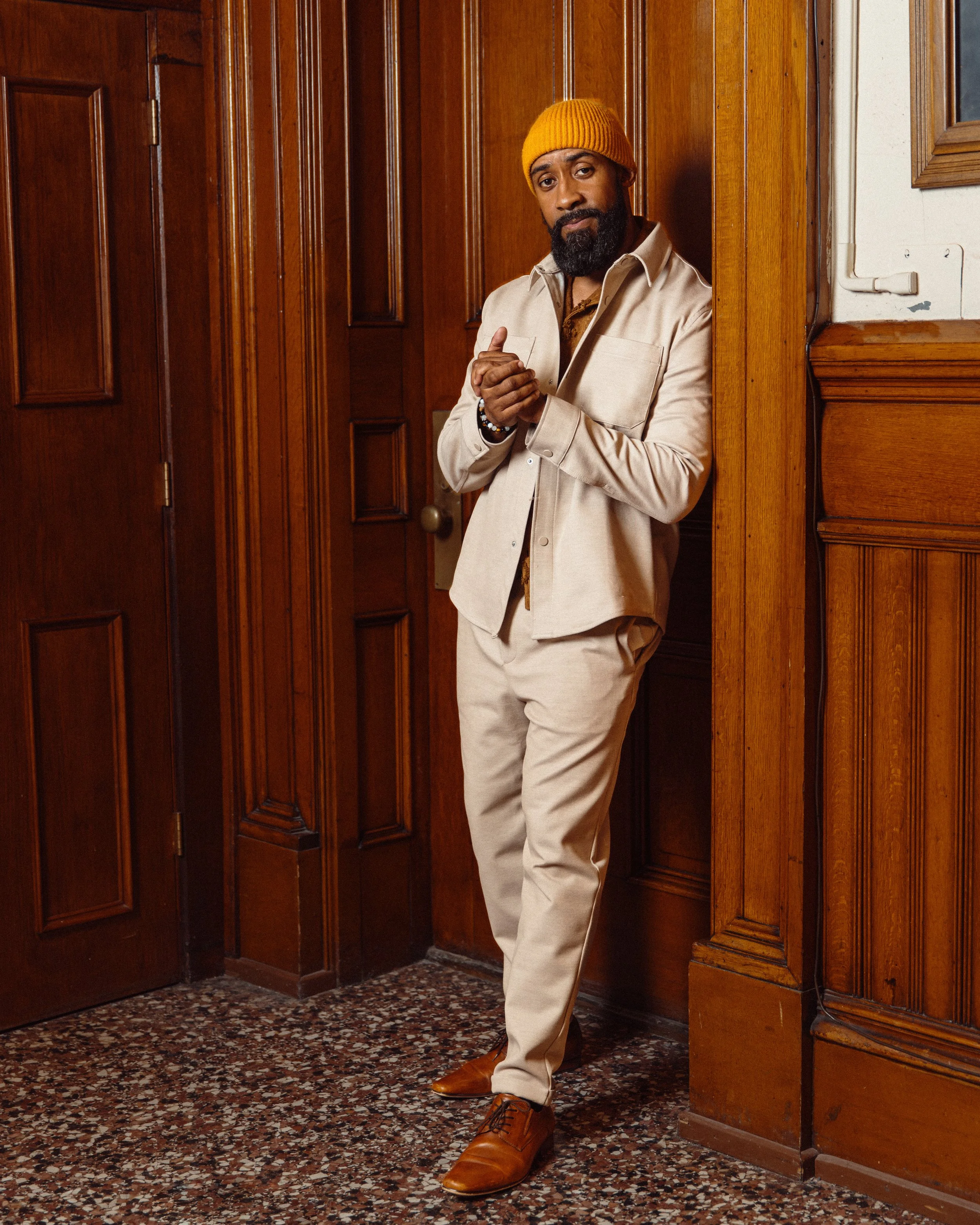 Rui Lopes of Anawan Studios in a beige suit standing in a room with wooden walls and patterned marble flooring, wearing a yellow knit beanie and brown shoes.