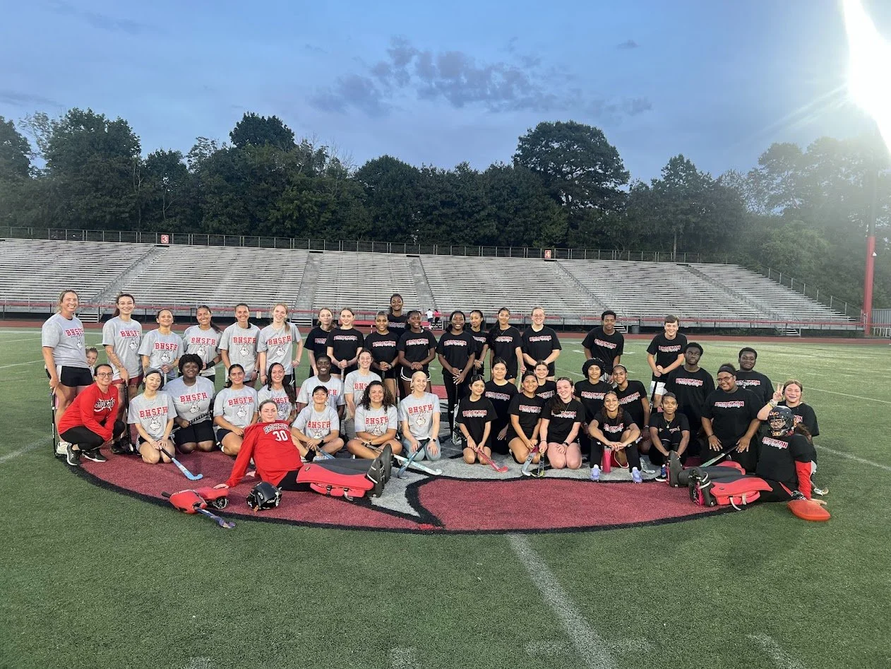 Group of girls and women posing with field hockey sticks on an outdoor field at a high school stadium during evening.