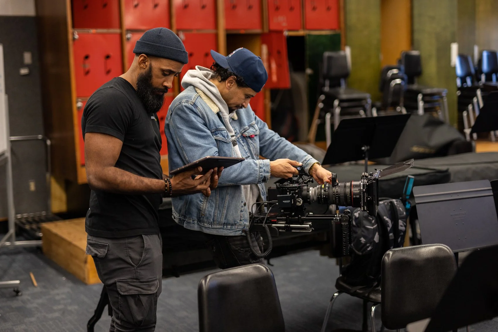 Two men in a room with red lockers and chairs, one adjusting a professional camera, the other holding a tablet.