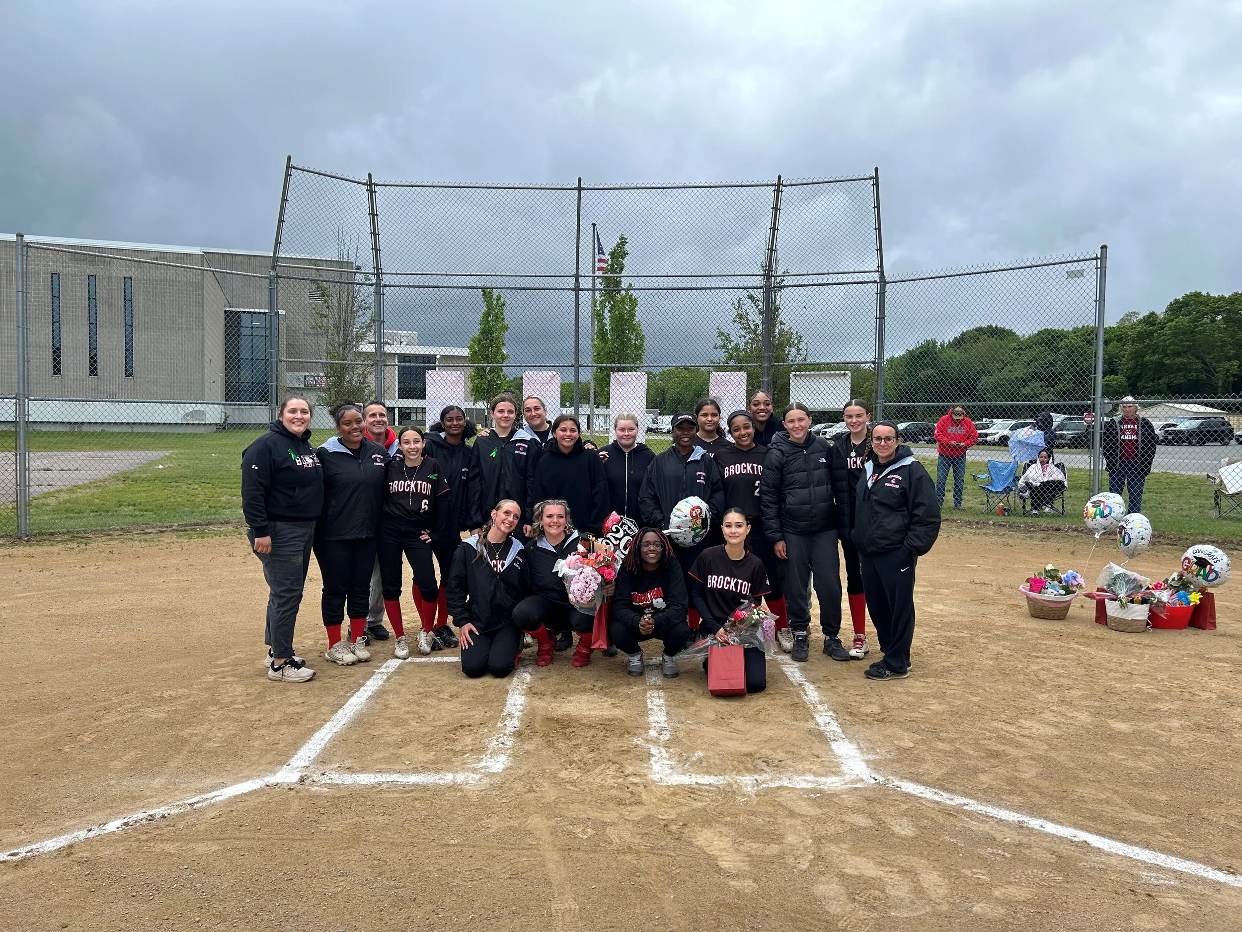 A group of women and girls in softball uniforms gathers on a baseball field for a photo, with some holding flowers. There are spectators and a few balloons in the background. The sky is overcast.