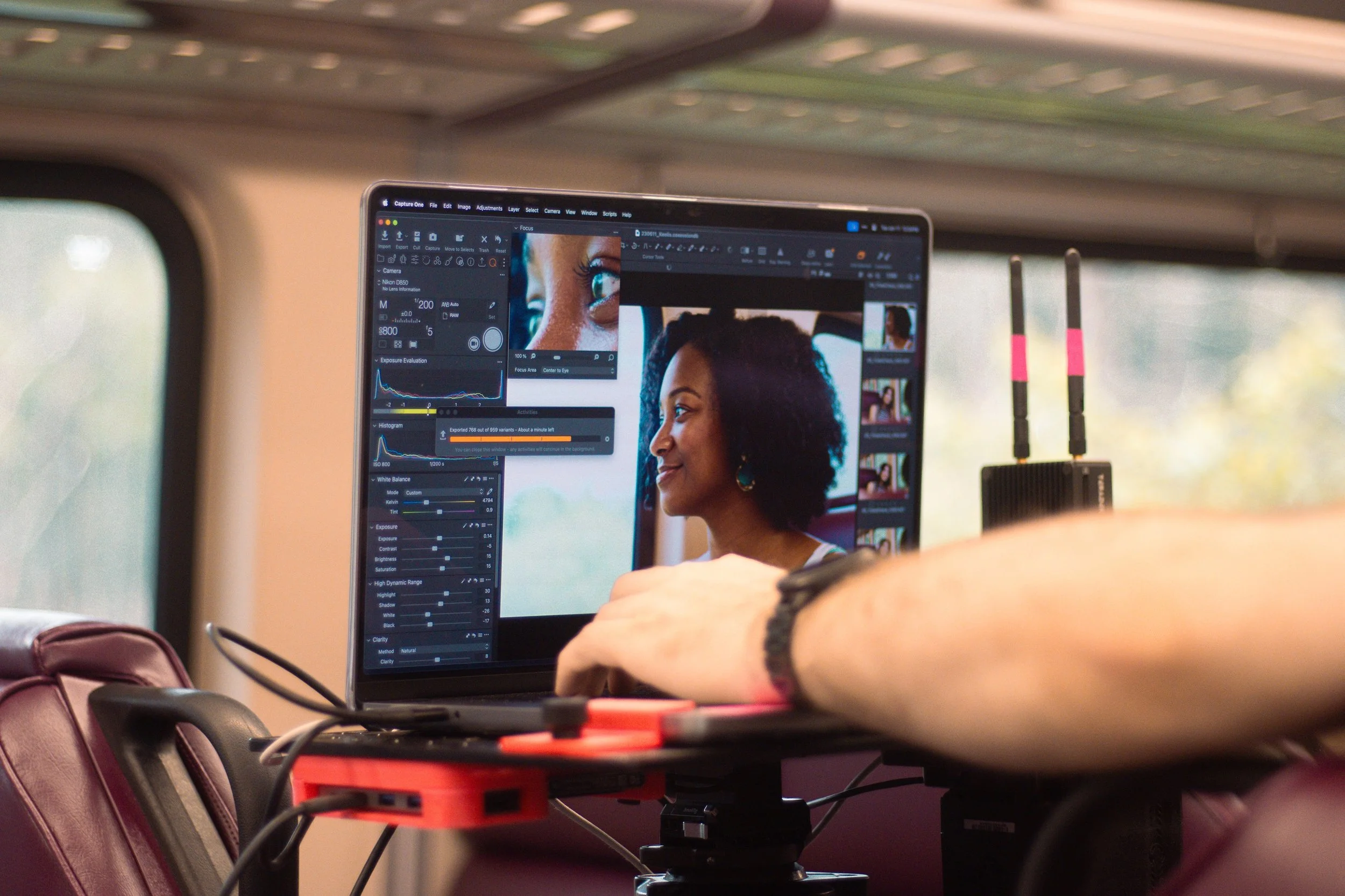 A close-up of a computer monitor displaying photo editing software with a woman with curly hair on the screen. The monitor is placed on a table with various equipment and cords around it. The setting appears to be a mobile or outdoor workspace, possi