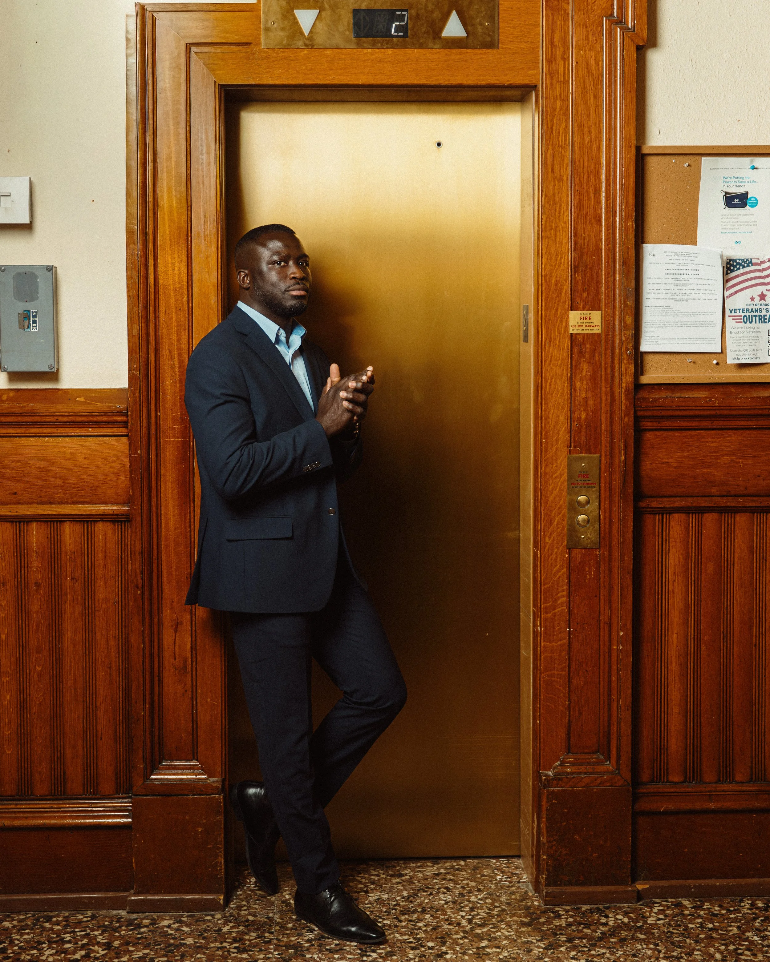 A man in a dark suit and light blue shirt stands inside an elevator with wooden paneling. He is leaning against the wall near the closed elevator door, looking at the camera, with one foot crossed over the other. The elevator has a digital display sh