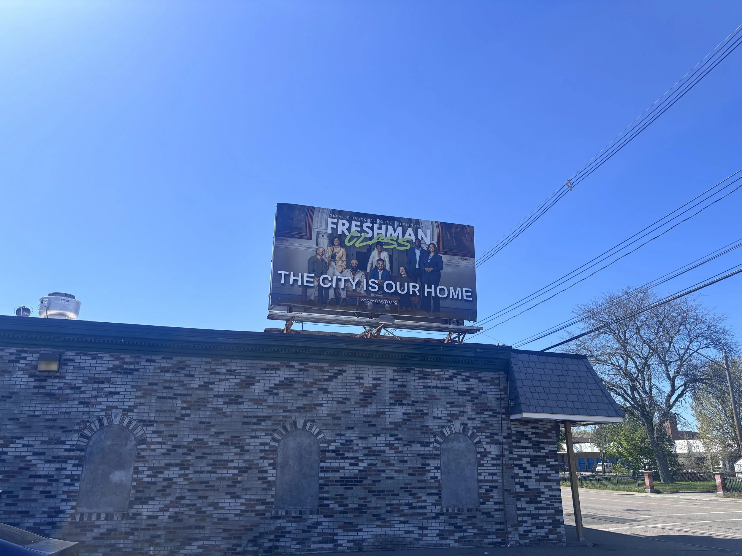 Billboard poster for 'FRESHMAN CLASS' with group photo of diverse young adults and the text 'THE CITY IS OUR HOME' at the bottom, mounted above brick building with two circular windows. Brockton MA