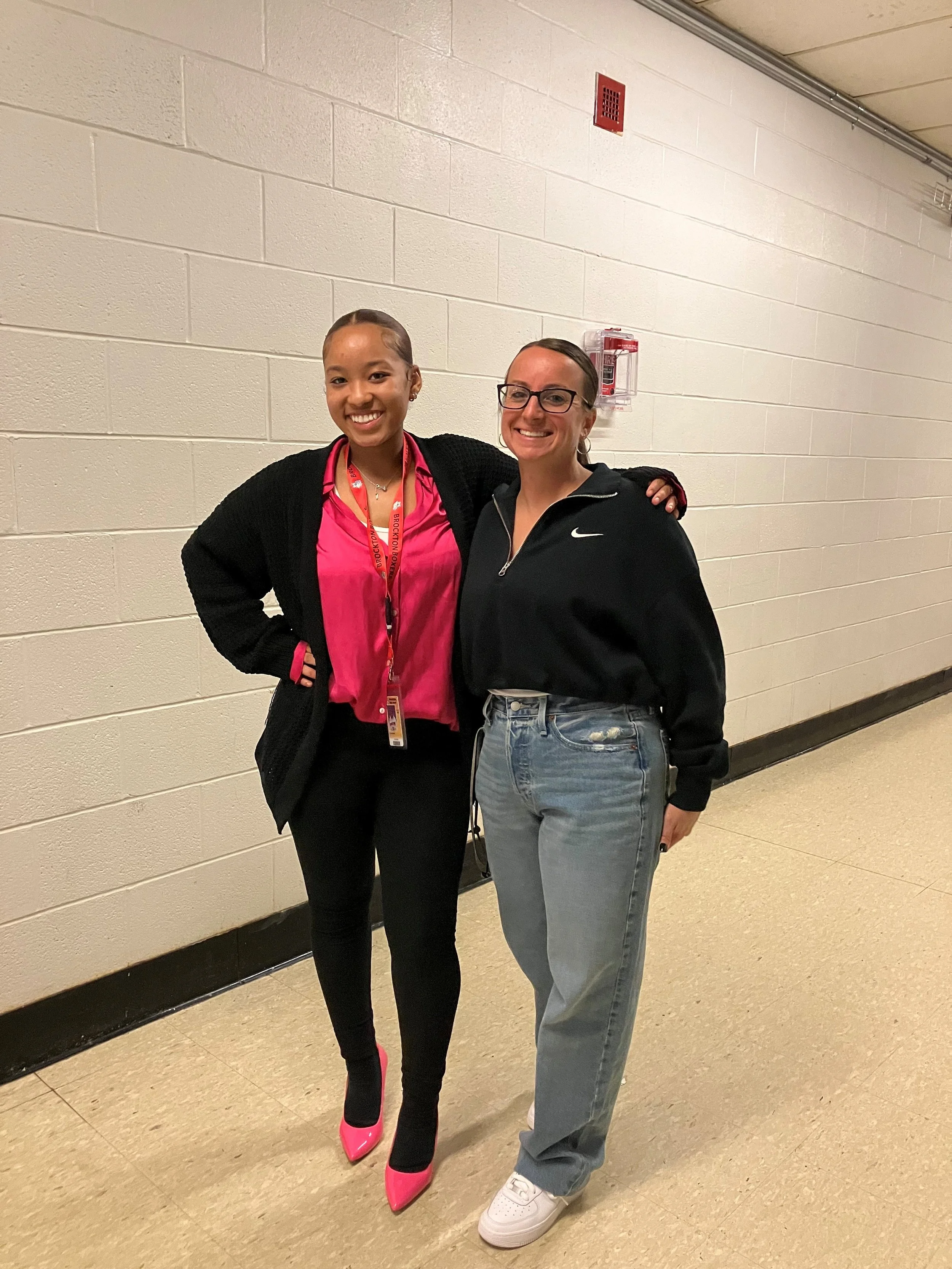 Two women standing close together in a hallway, smiling at the camera. The woman on the left has dark skin, is wearing a pink blouse, black cardigan, black pants, and pink high-heeled shoes. She has dark hair pulled back and is wearing a lanyard. The