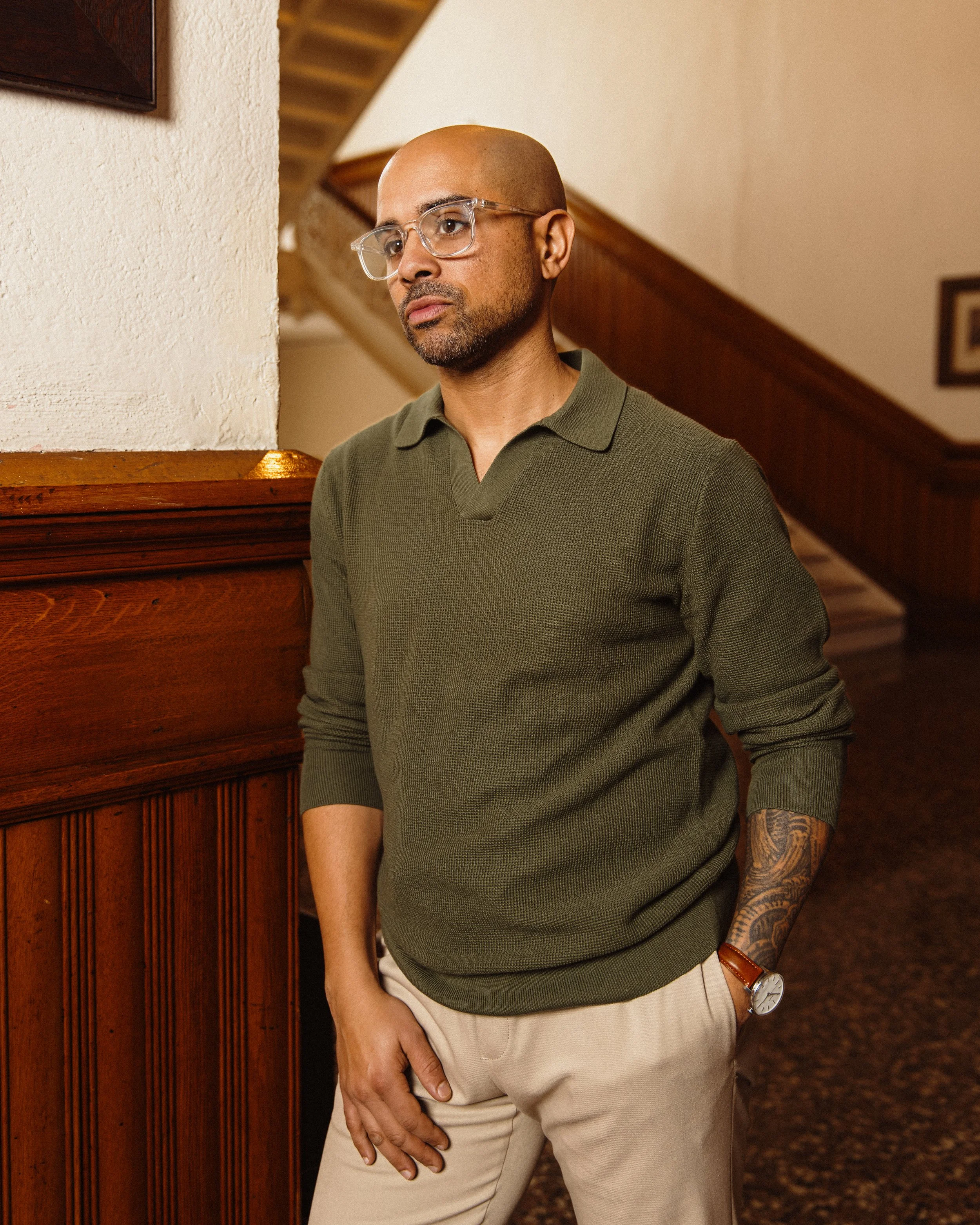 Nelson Fernandes wearing glasses, a green sweater, and beige pants standing indoors next to a wooden wall with stairs in the background.