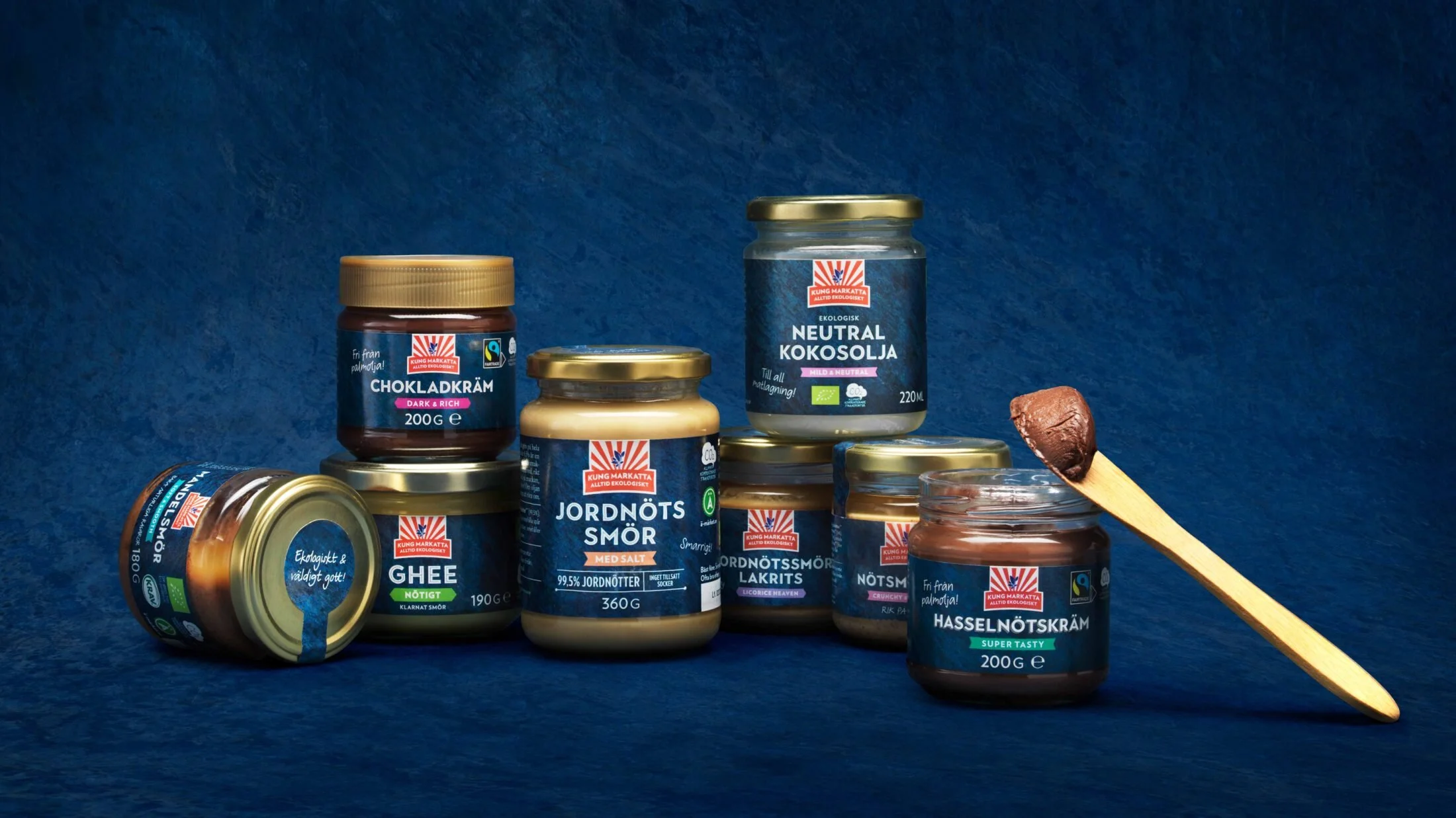 Arranged jars and cans of organic spread, ghee, and nut cream on a blue background with a wooden spatula