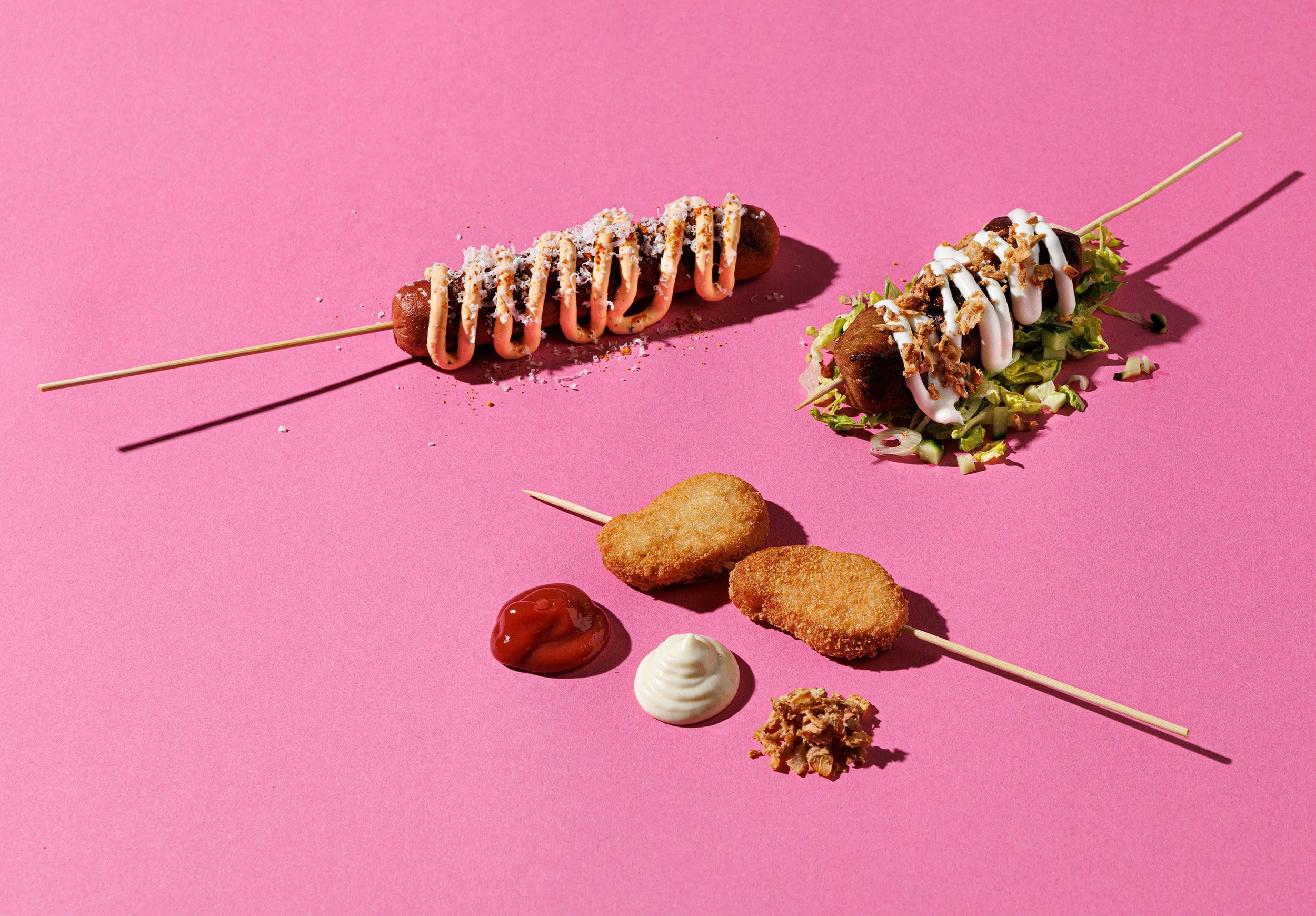Three skewered foods with various toppings and sauces on a pink background, including a hot dog with drizzle, a salad with dressing, and fried chicken nuggets with dipping sauces.