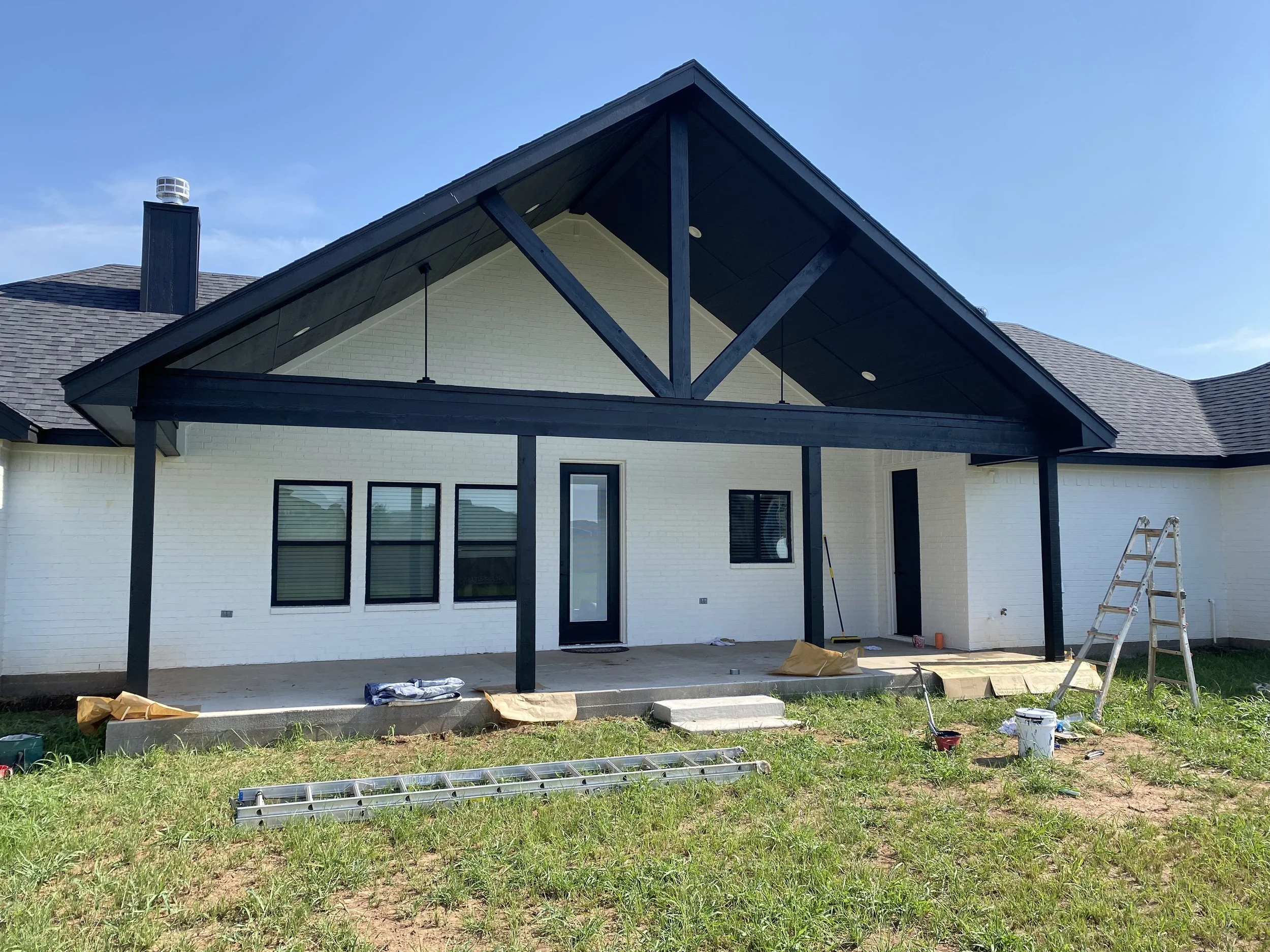 Rear view of a house under construction with a large covered patio and black wooden beams. The exterior is white brick with several windows and a door. A ladder and construction materials are visible on the grass.