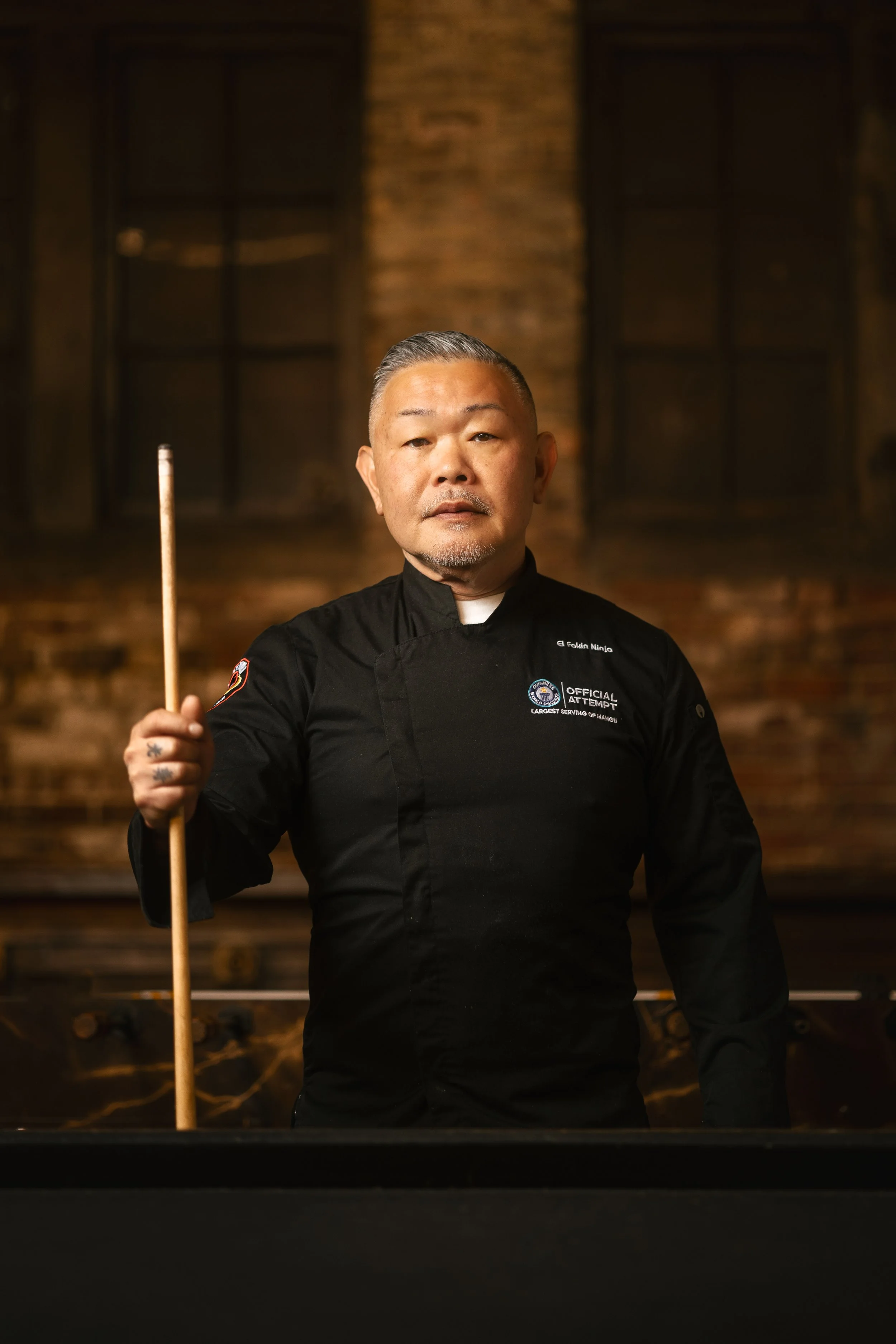 A professional chef dressed in black uniforms with patches, holding a pool cue, standing in a dimly lit bar or club with brick walls in the background.