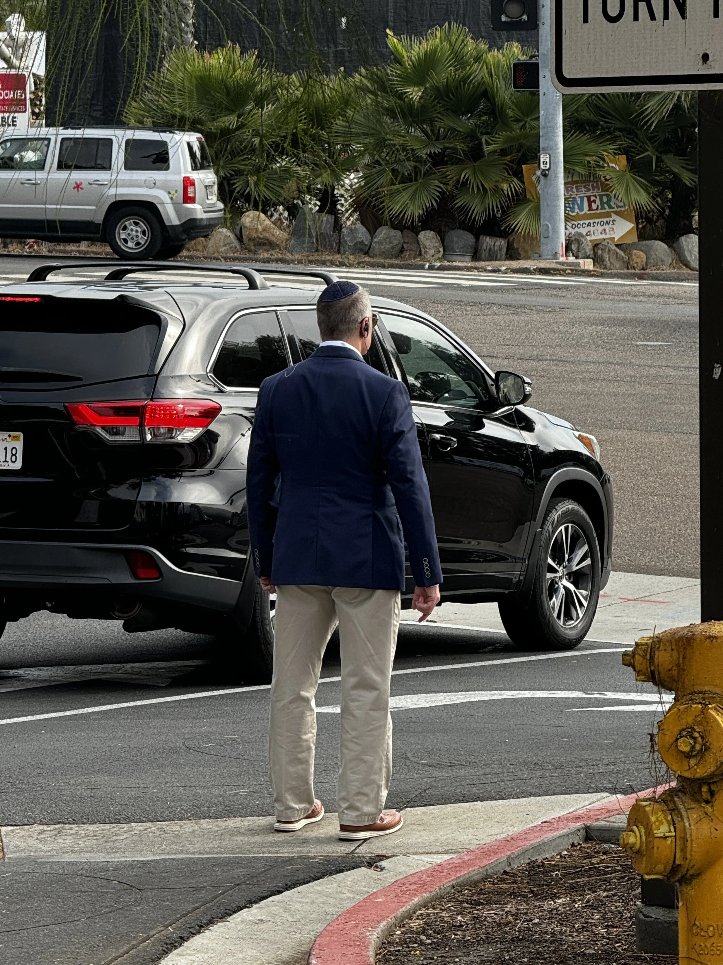 A man wearing a navy blazer, beige pants, sandals, sunglasses, and a kippah stands next to a black car on a city street. There is a fire hydrant in the foreground and palm bushes across the street.
