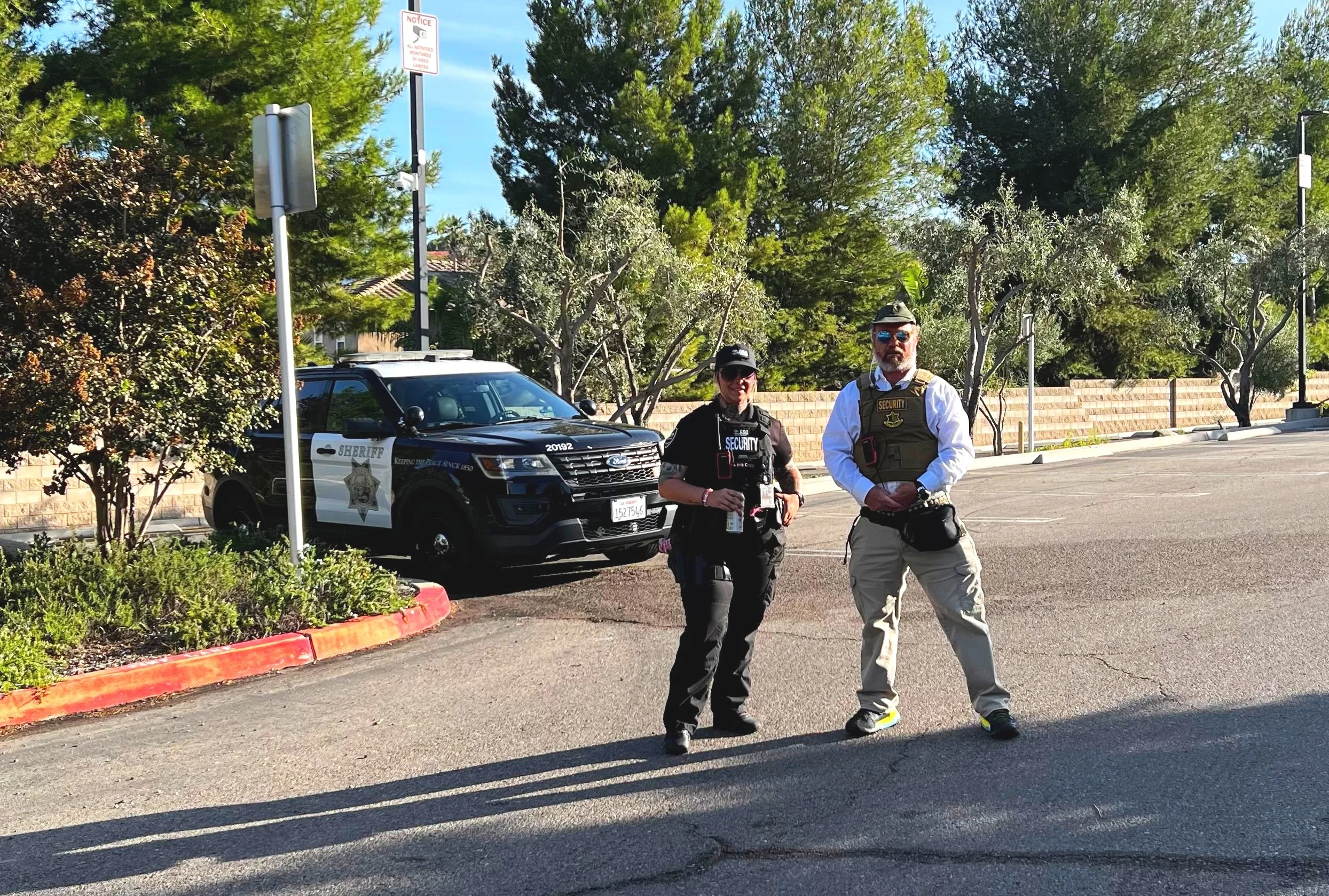 Two security personnel, one woman and one man, standing on an asphalt parking lot near a sheriff's vehicle with trees and a brick wall in the background.