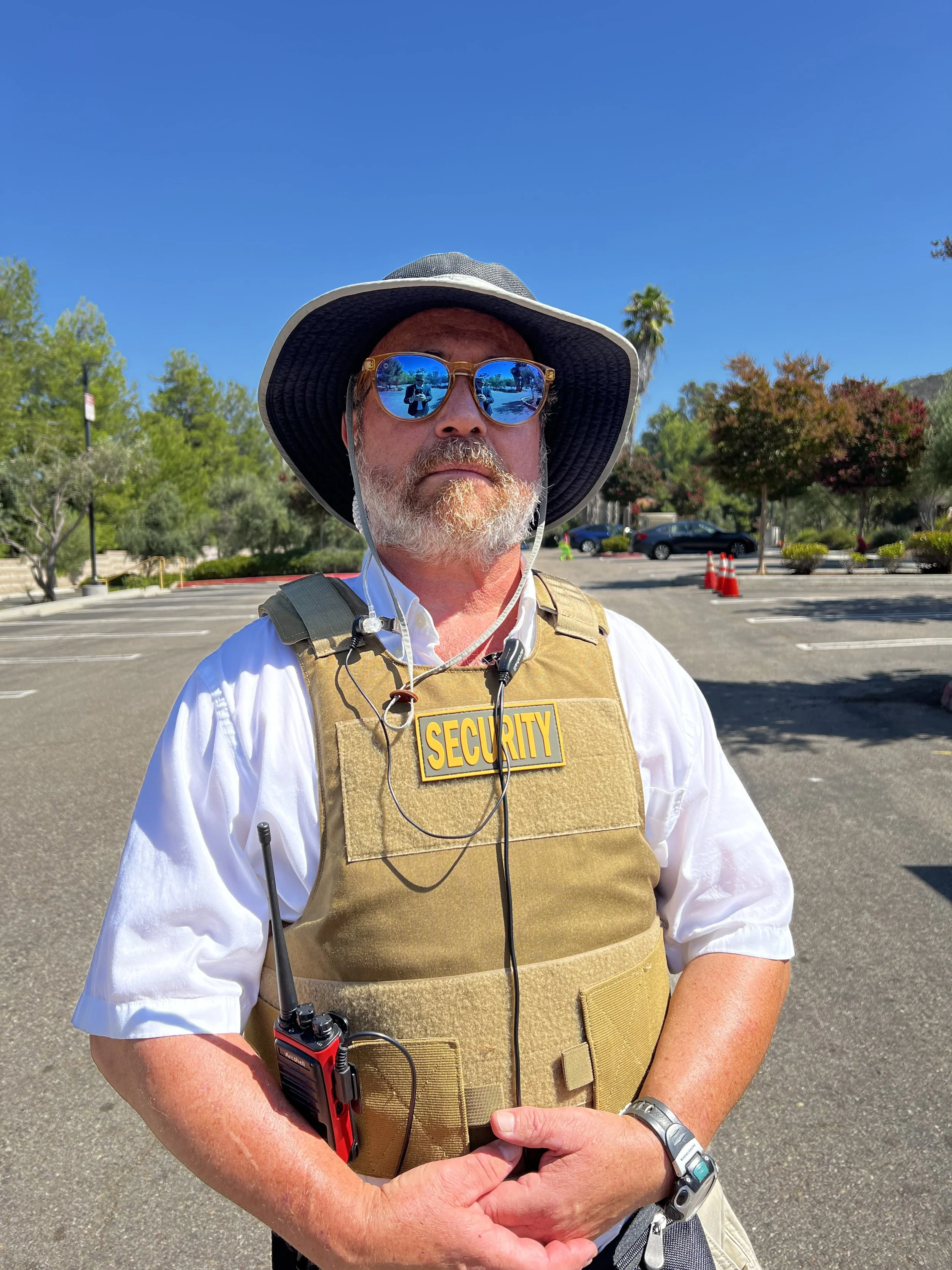 Security guard wearing a tan vest with a 'Security' badge, sunglasses, a wide-brimmed hat, and a watch, standing outdoors in a parking lot under bright sunlight.