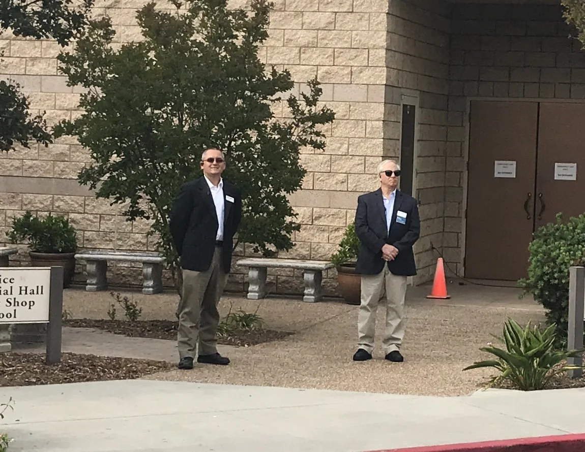 Two men in business attire with sunglasses standing outside near a brick building, with potted plants and a sign that reads 'Police,' 'Town Hall,' and 'Shop'.