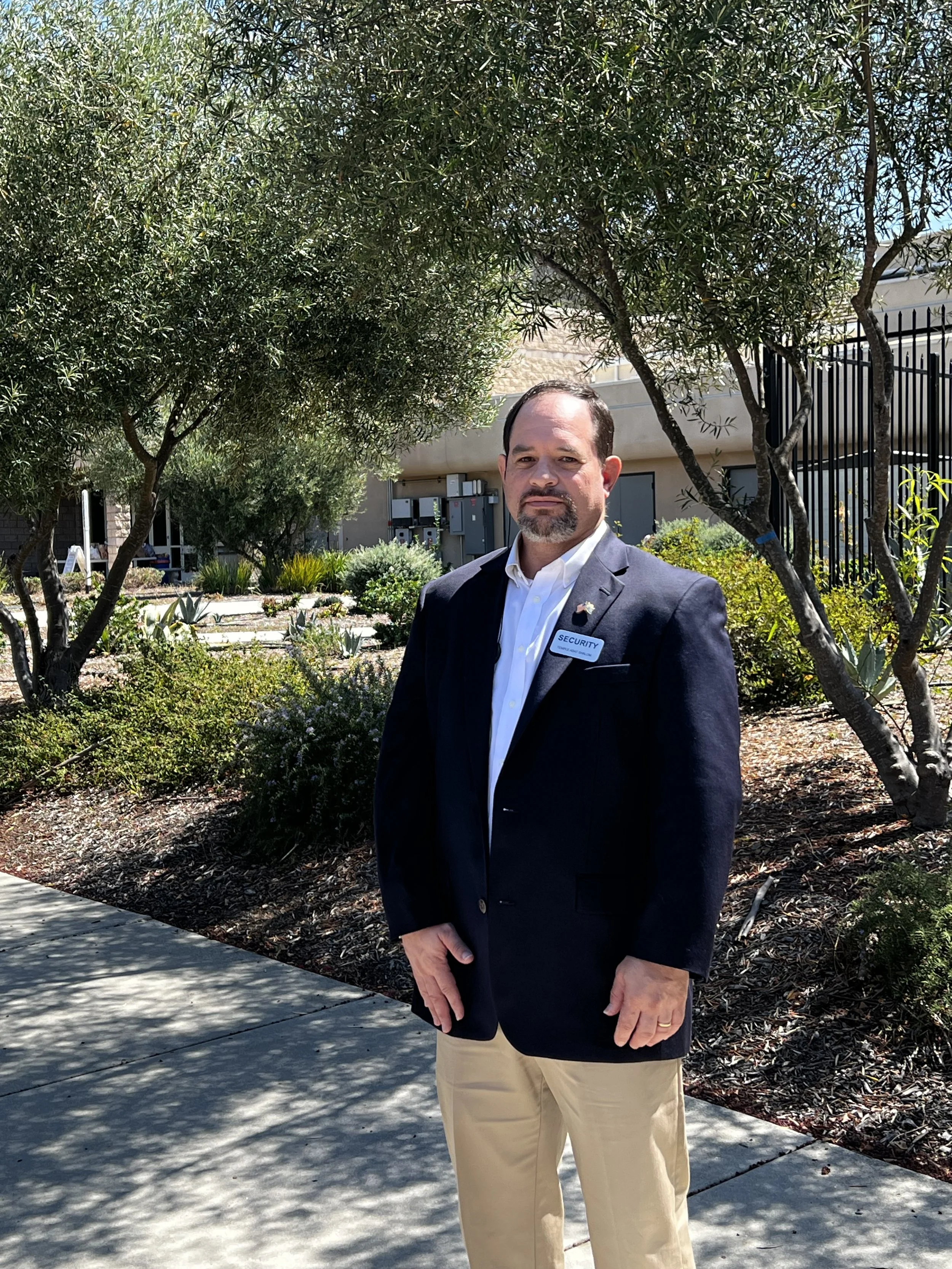 A man in a black blazer with a security badge standing outdoors on a sunny day among trees and plants.