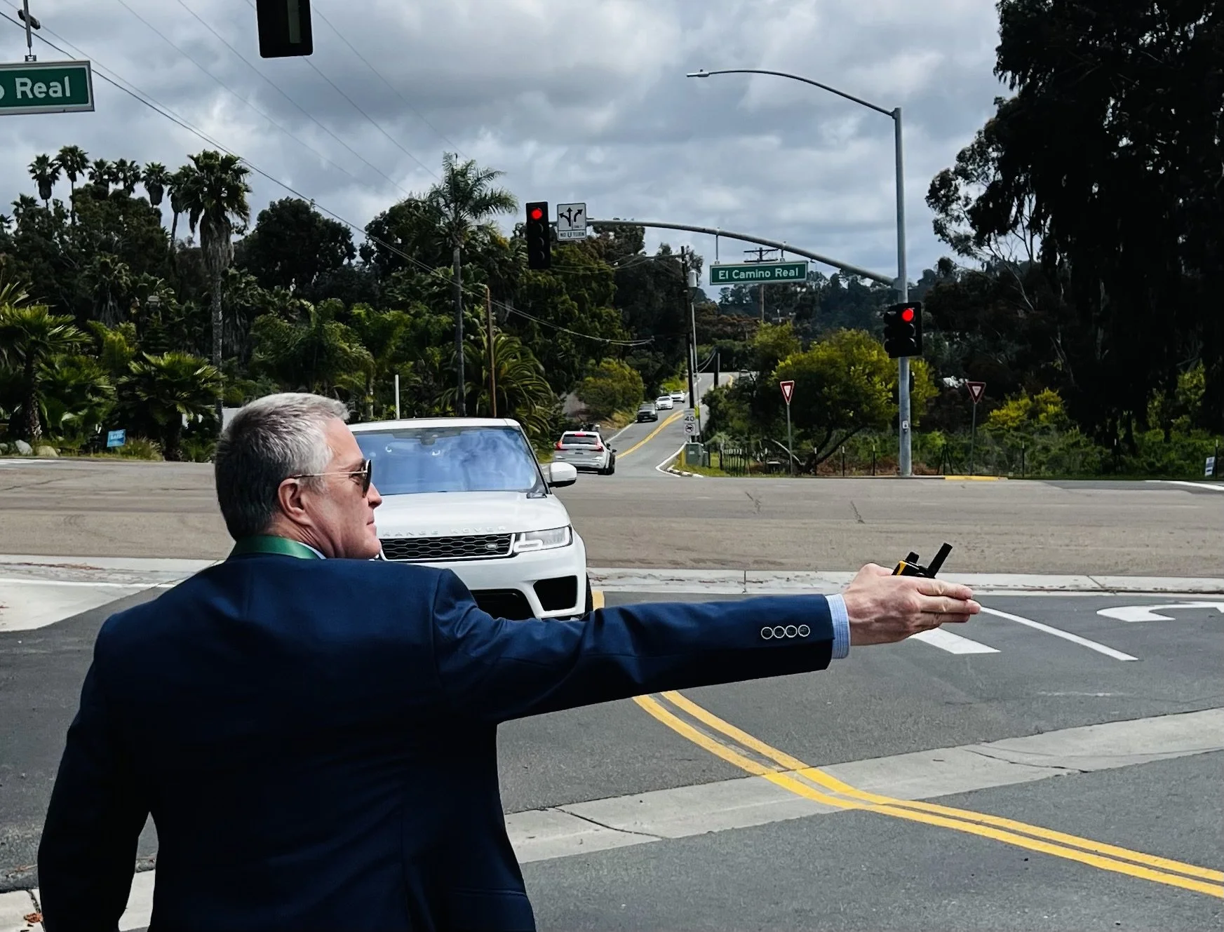 A man in a dark suit with gray hair is standing at an intersection, holding a remote control in his outstretched hand, with traffic lights and cars in the background under a cloudy sky.
