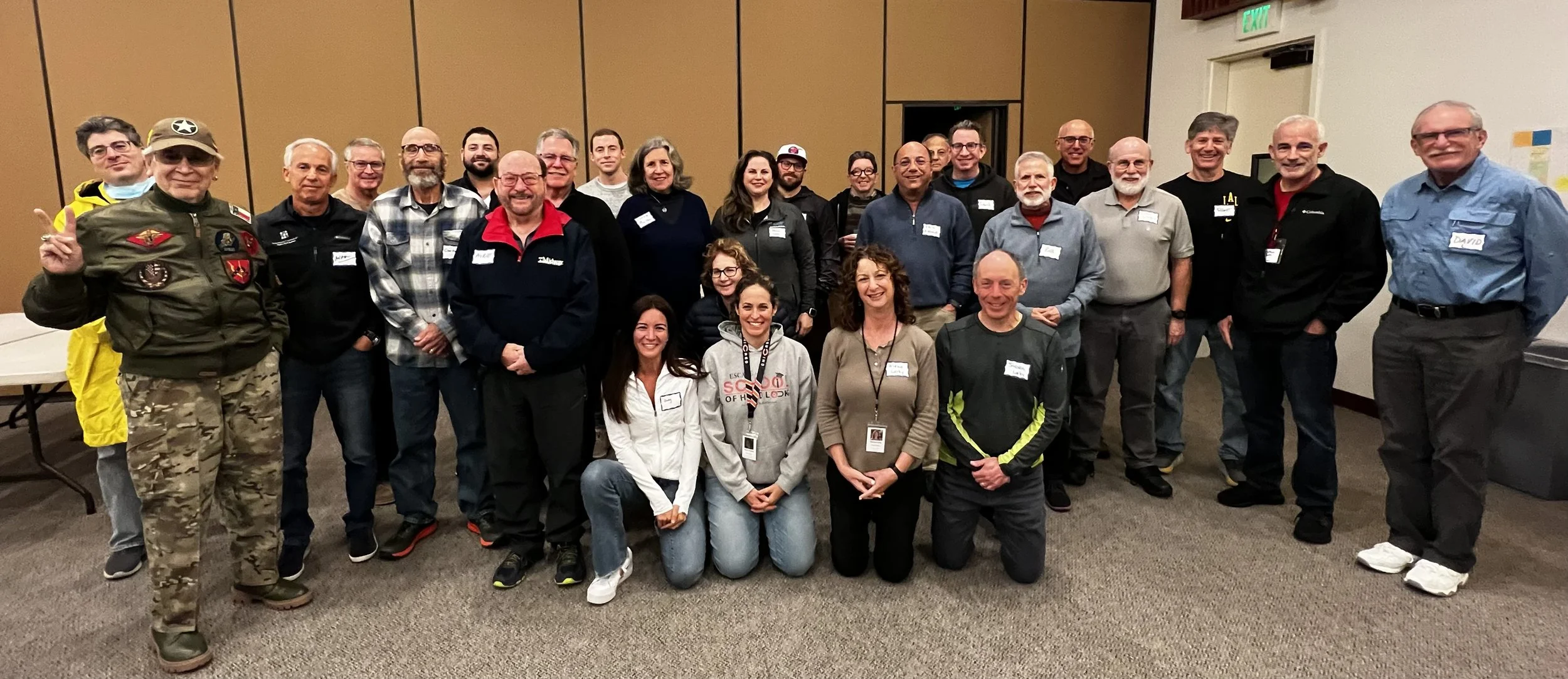 Group of diverse adults posing indoors for a photo, some with name tags, in a room with brown and white walls.