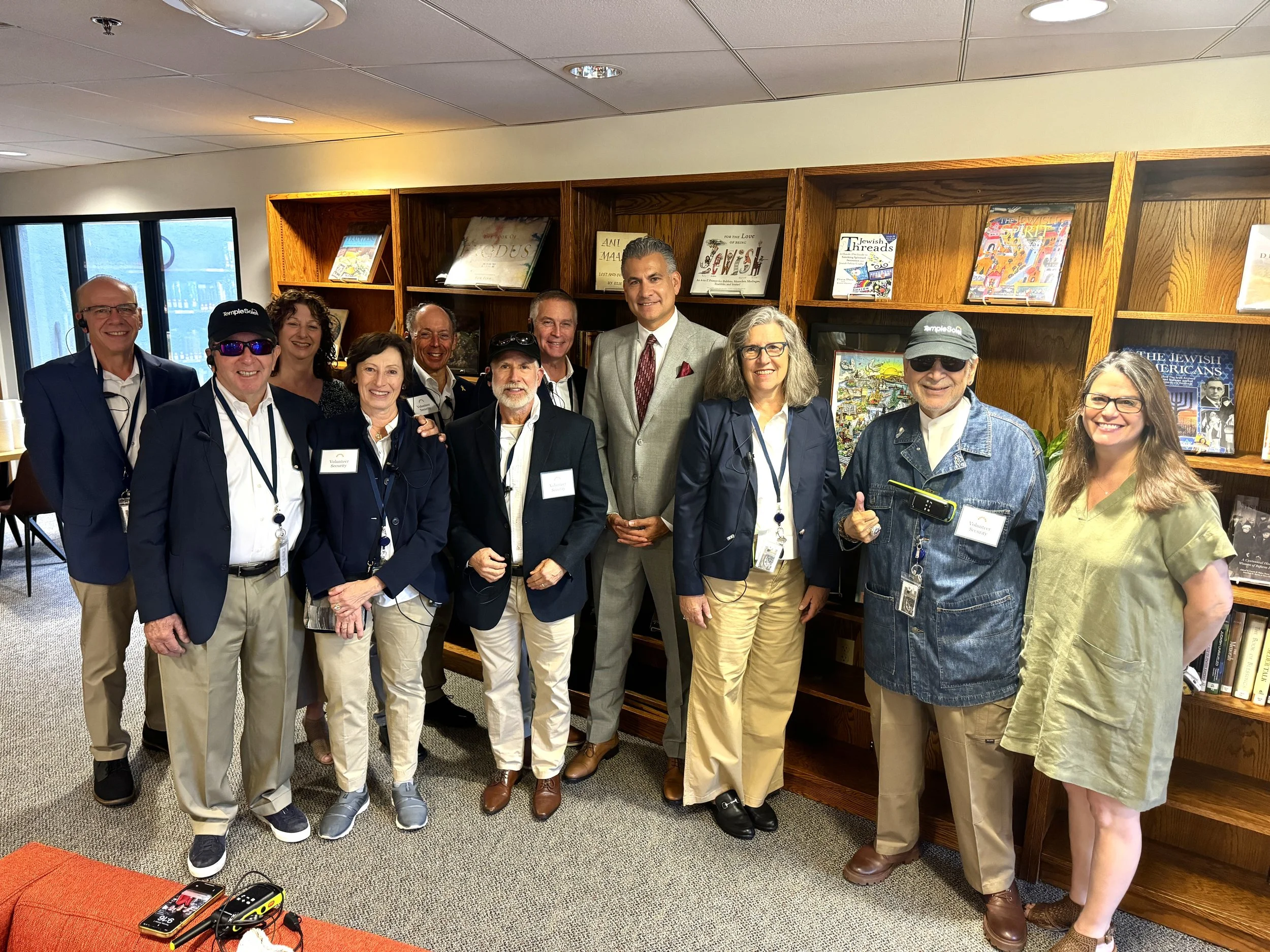 Group of eleven people posing indoors near a wooden bookshelf with books and artwork, with a window on the left and a table with electronic devices in the foreground.