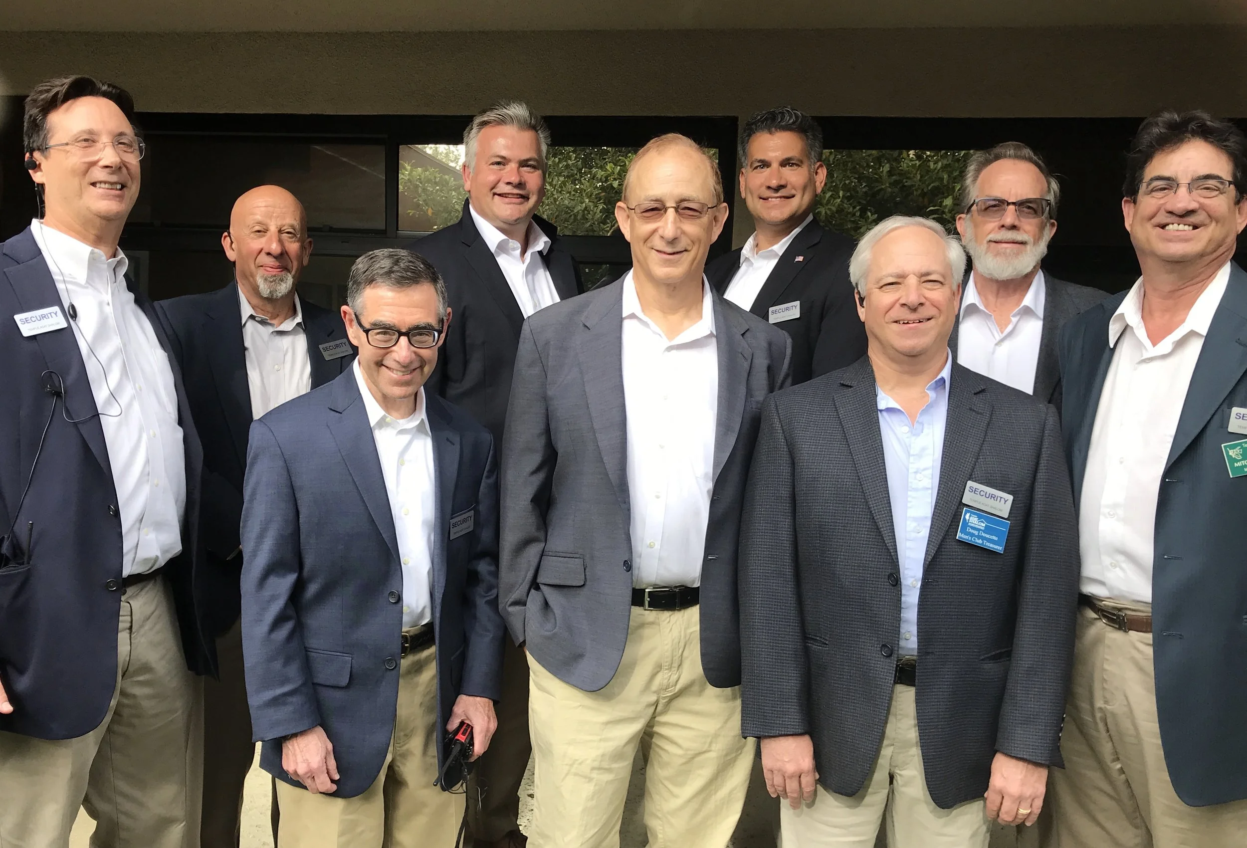 Group of men in business suits gathered together outdoors for a photo, most wearing name tags.