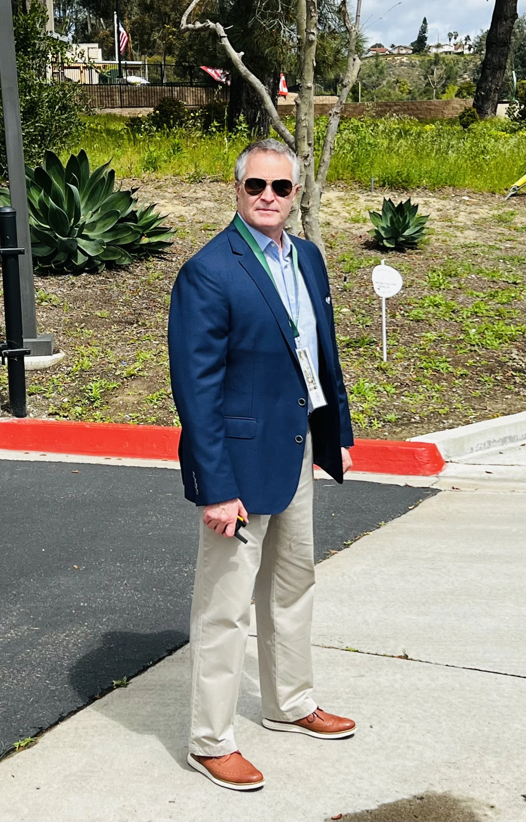 A man wearing sunglasses, a blue blazer, a light blue shirt, beige pants, and brown shoes standing on a sidewalk near a landscaped area with succulents and trees.