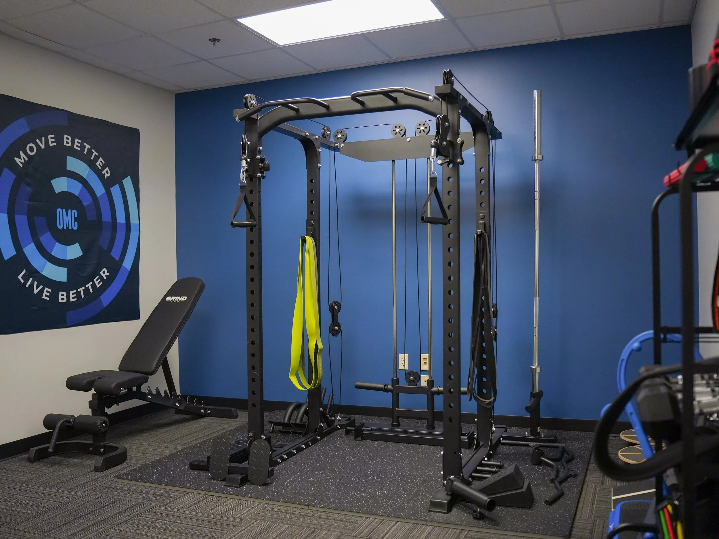 A compact workout space with a black weightlifting rack and a bench, resistance bands hanging from the rack, and a poster with blue concentric circles and the words 'Move Better, Live Better' on a white wall. The room has blue and white walls and dark gray carpet flooring.