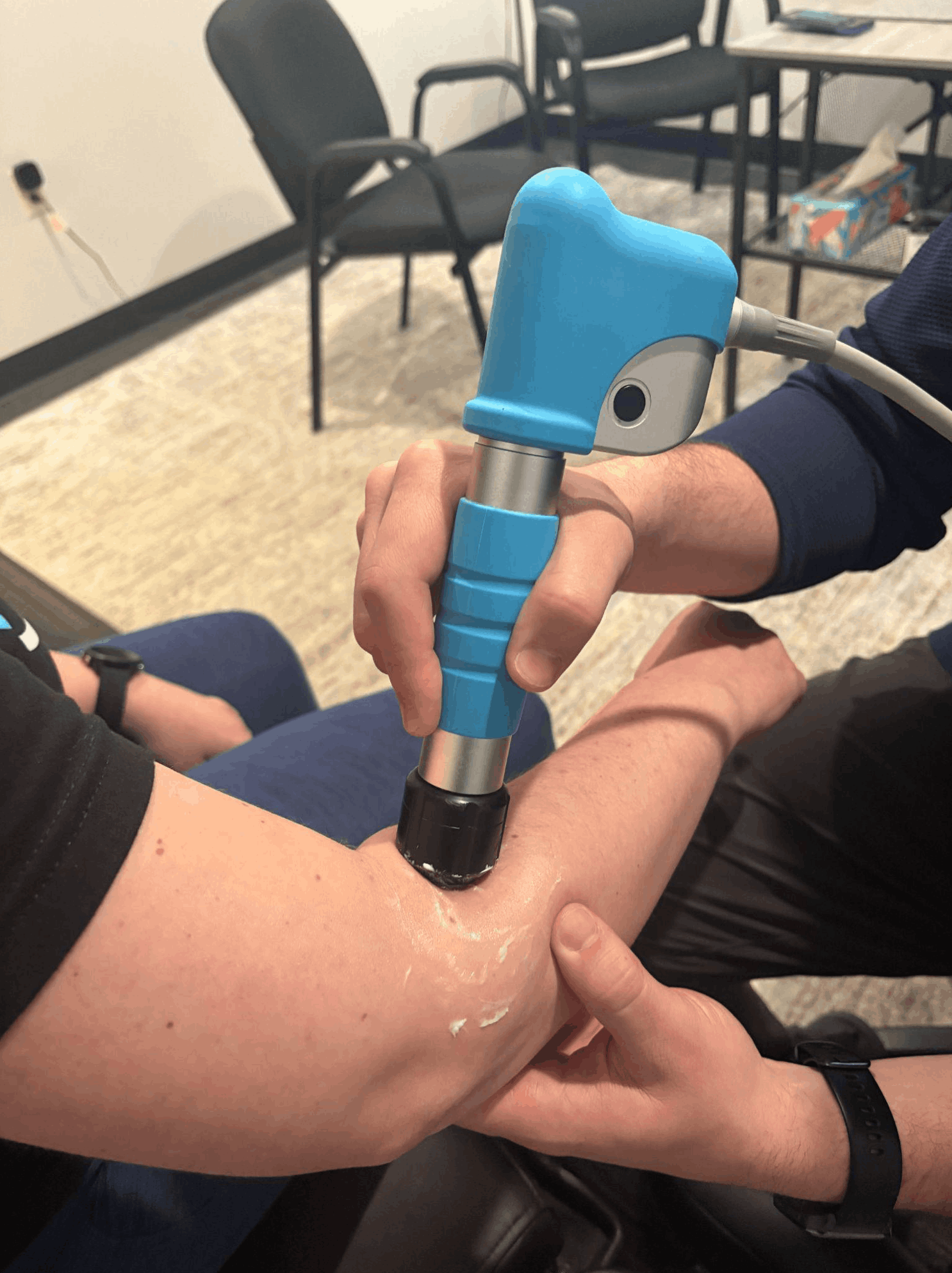 Person receiving physical therapy or a medical treatment involving a handheld device on their leg, in a room with chairs and a table, and a box of tissues in the background.