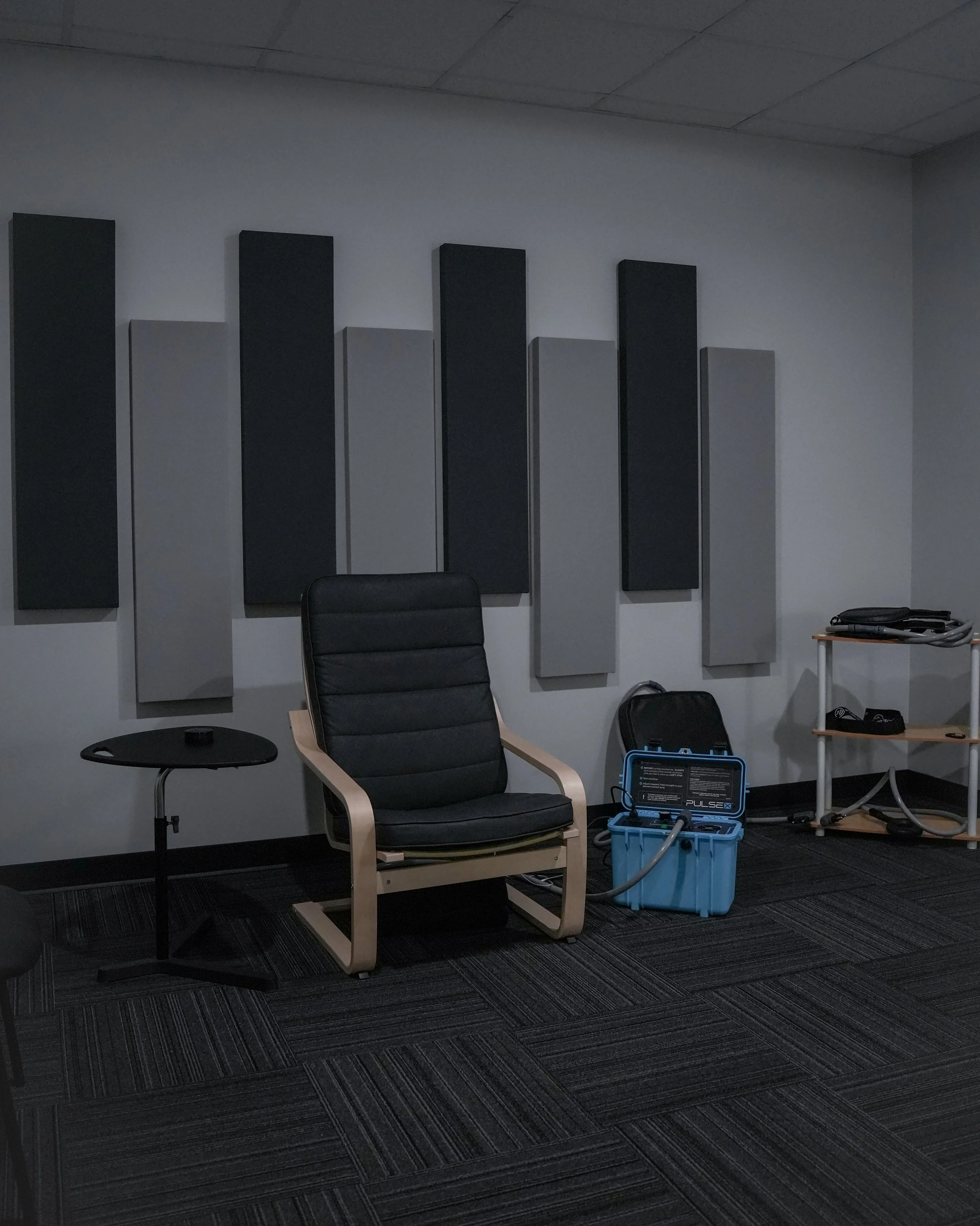 An office or conference room with acoustic wall panels, a black office chair, a small black side table with a remote on it, and a treadmill or exercise machine in the corner with a blue toolbox and black cords on the floor.