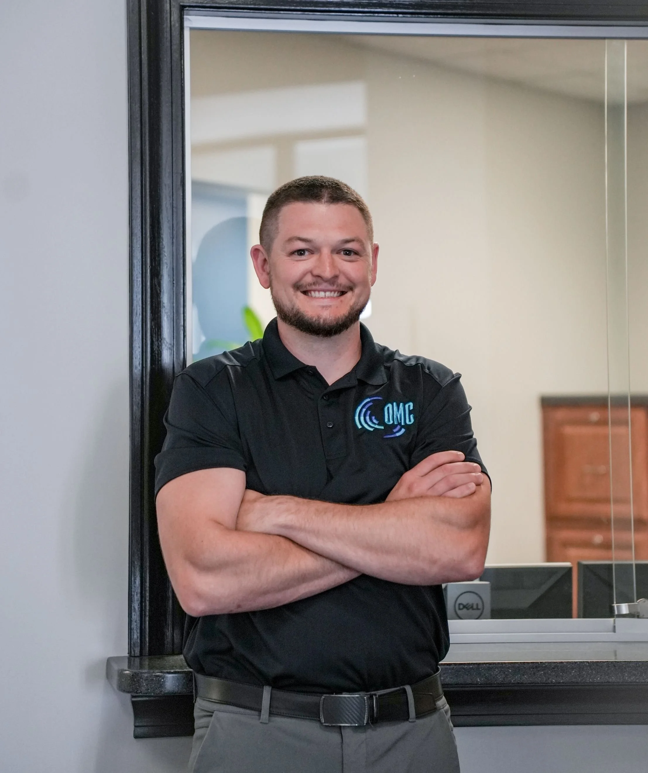 A man with short hair and a beard, smiling, standing with arms crossed in front of a window and classroom. He is wearing a black polo shirt with a logo and gray pants with a belt.