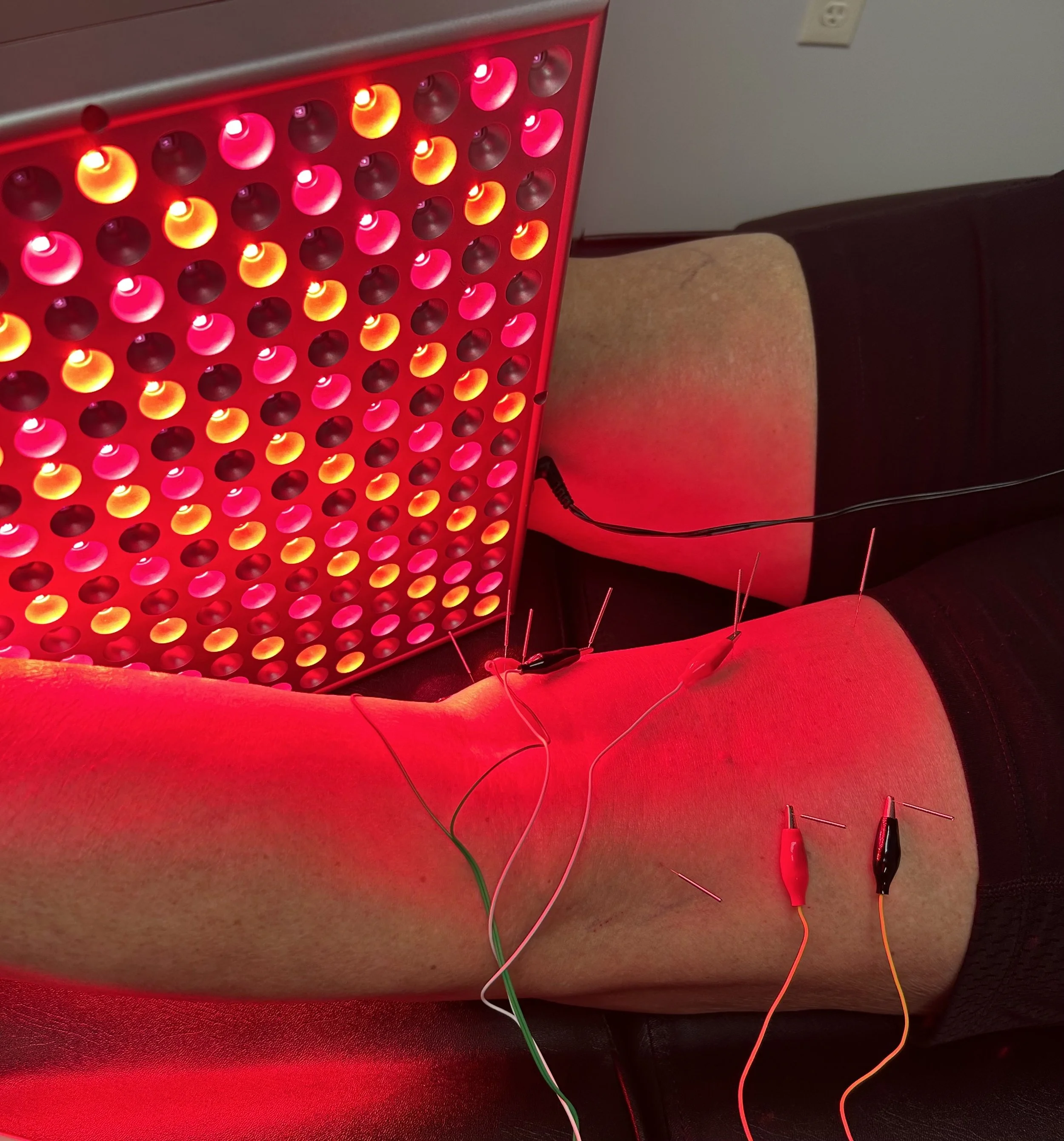 Close-up view of an electronic device with multiple orange and pink LED lights in a grid pattern, with wires connected to it, positioned on a red seat in a room with a beige and black seat and a wall with an electrical outlet.