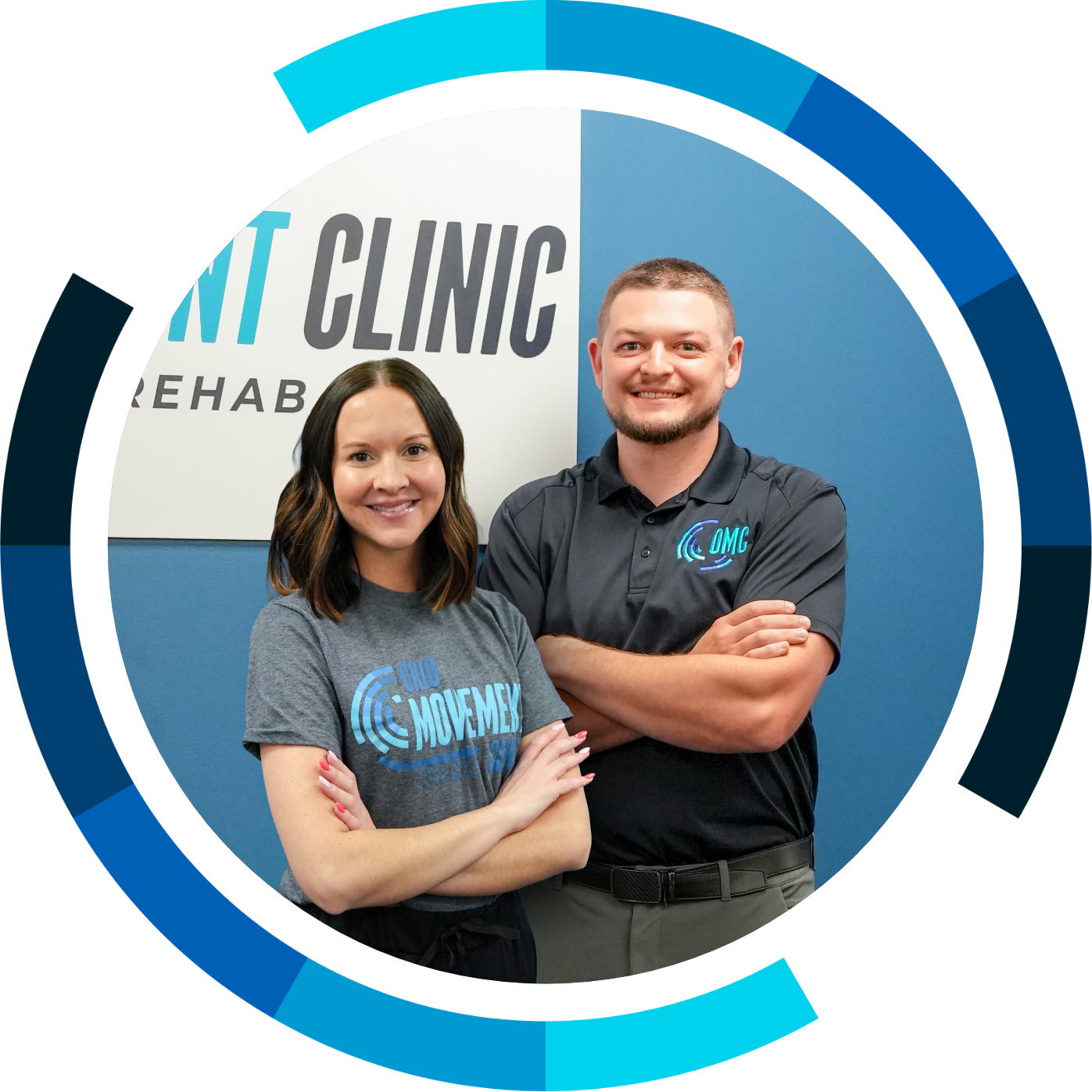 A woman and a man smiling and standing with arms crossed in front of a sign that says 'REHAB CLINIC.' The woman is wearing a gray T-shirt with 'OMG MOVEMENT' on it, and the man is wearing a black polo shirt with the same logo.