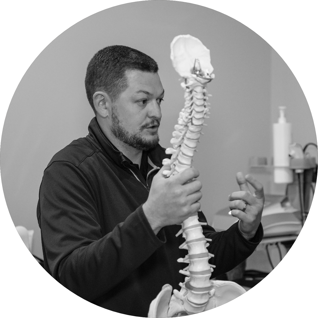 A man with short hair and a beard holding a model of a human spine, explaining it in a medical or educational setting.