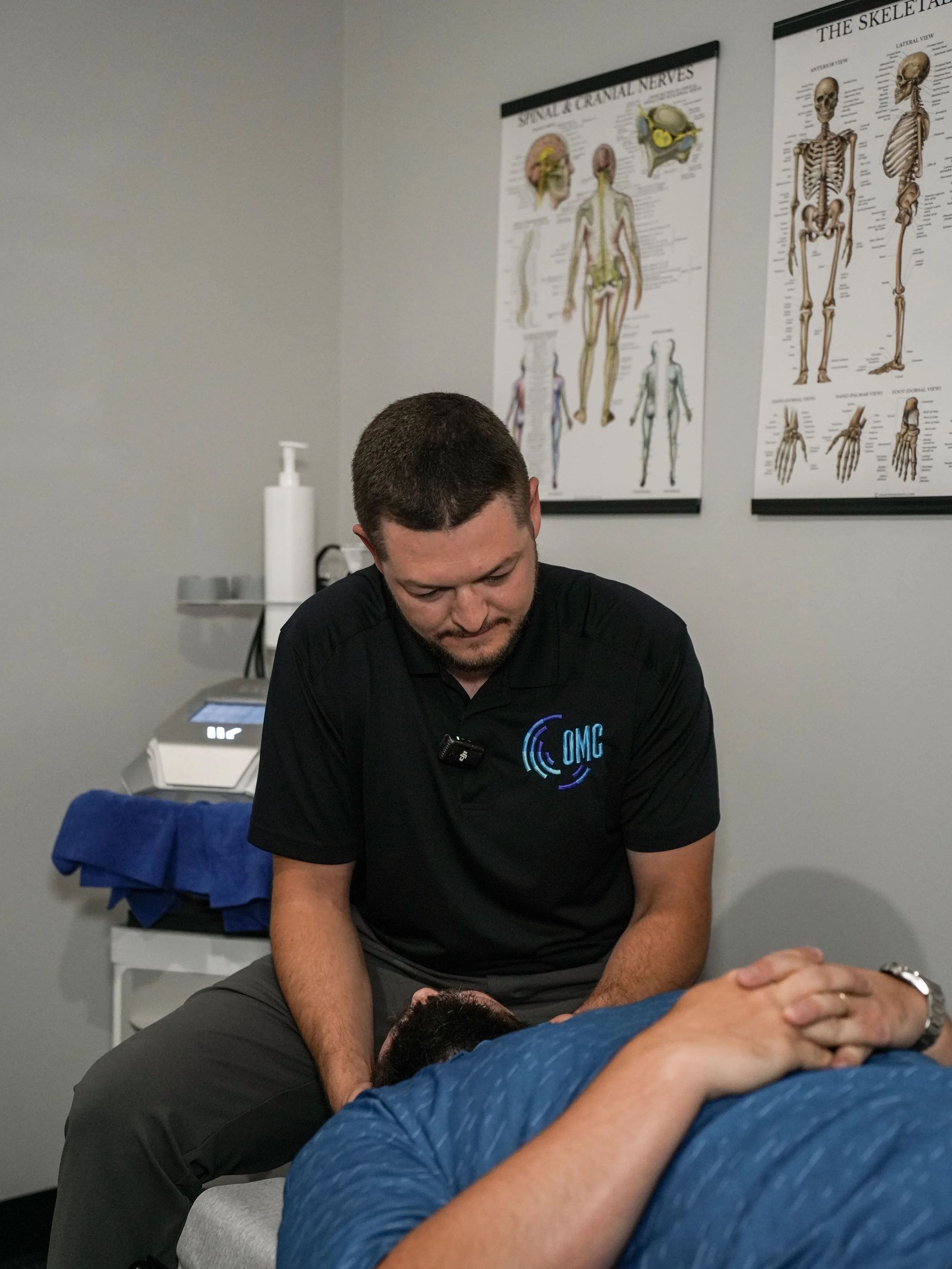 A man in a black shirt performing a chiropractic adjustment on a patient lying face down on a table. The background features anatomy posters on the wall.