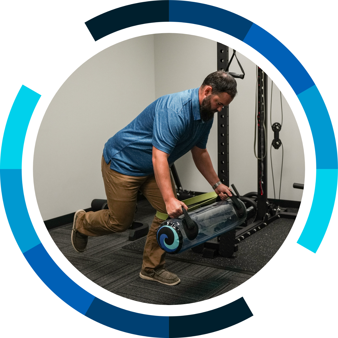 A man in a blue polo shirt and khaki pants performing a kneeling exercise with a water-filled workout barrel in a gym, surrounded by workout equipment and black rubber flooring.