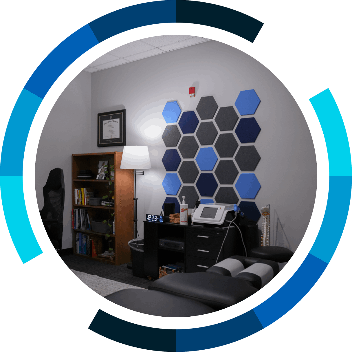 Office room with a black desk, a white medical device, a bookshelf, a lamp, and a wall decorated with hexagonal acoustic panels in shades of blue and gray.