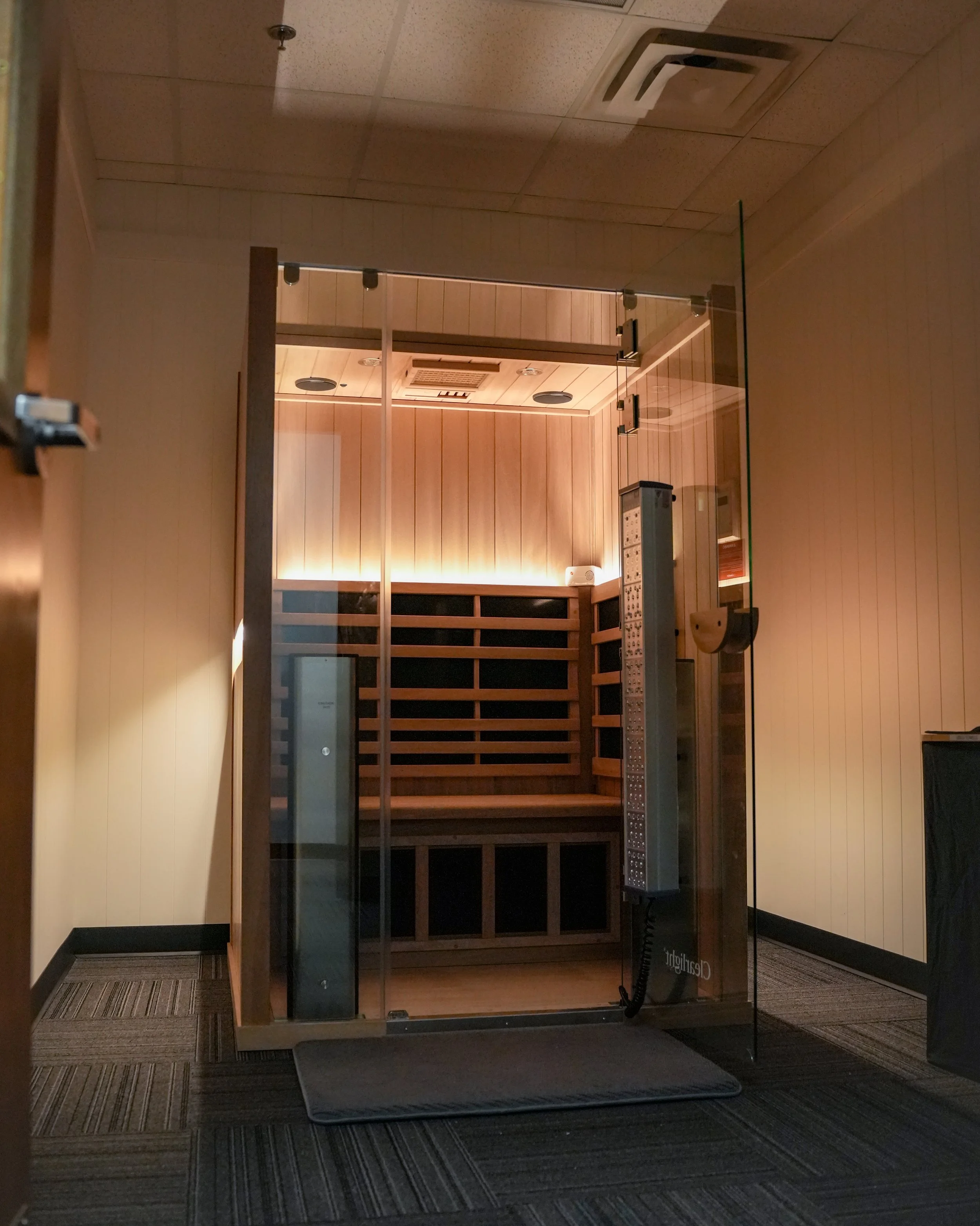 A wooden sauna room with a glass door, inside there are wooden benches and a sauna heater in the corner. The room has a wood-paneled ceiling with recessed lighting, and there is an air vent on the ceiling. The image is viewed from outside through a glass wall.