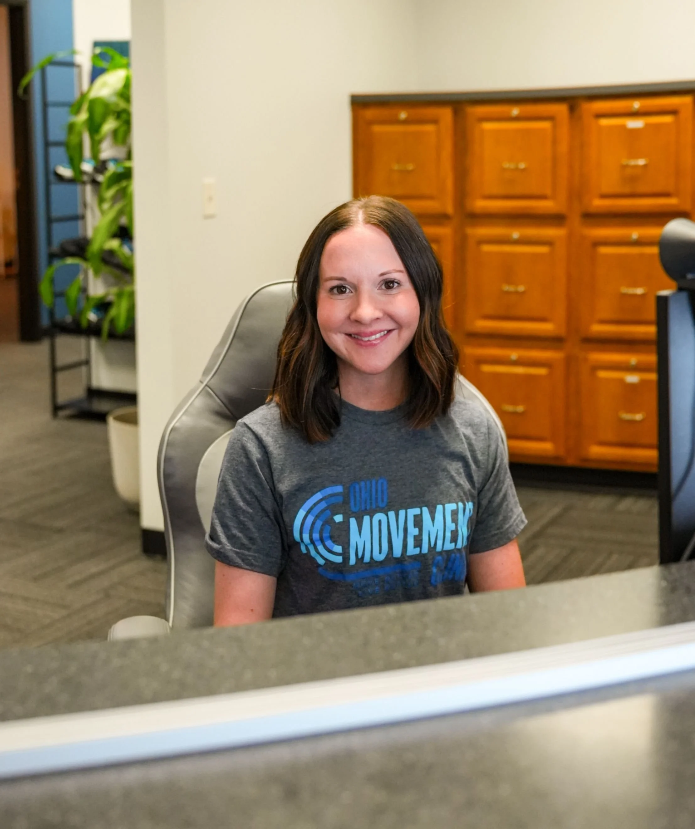 A woman with medium-length brown hair and fair skin, smiling, wearing a gray T-shirt with blue text, sitting at a desk in an office setting, reflected in a mirror or glass surface.