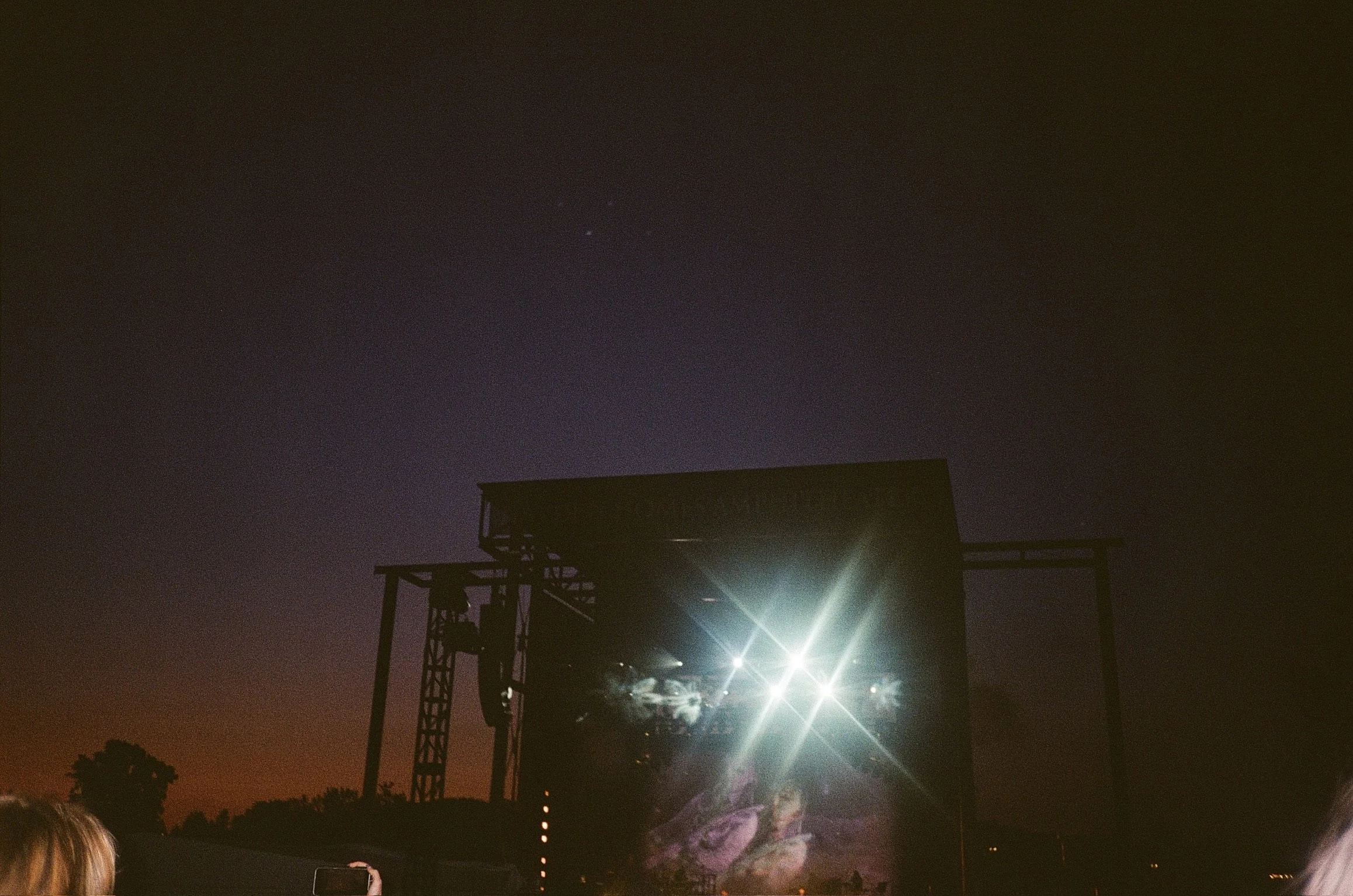 An outdoor concert stage lit up at night with bright lights, a large screen displaying visuals, and a dark sky overhead.