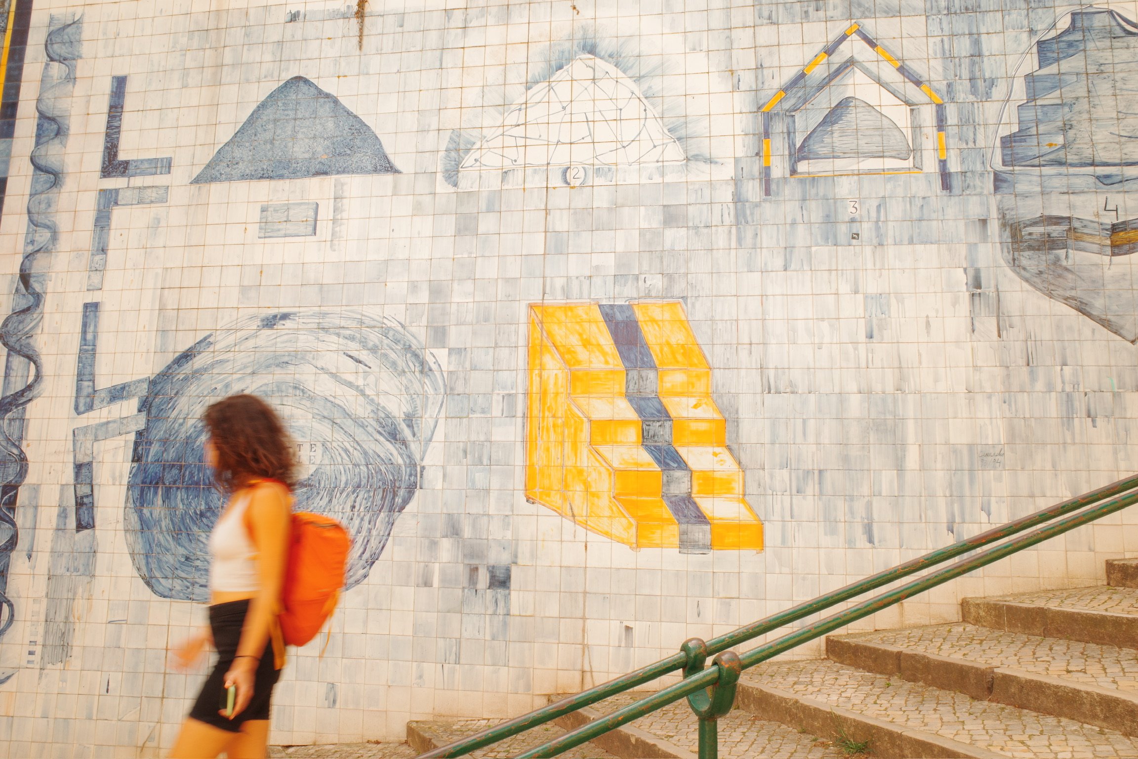 A woman with curly hair wearing a white top, black shorts, and an orange backpack walking in front of a large mural on a tiled wall. The mural features geometrical shapes and architectural sketches, including a house, a staircase, and a tunnel.