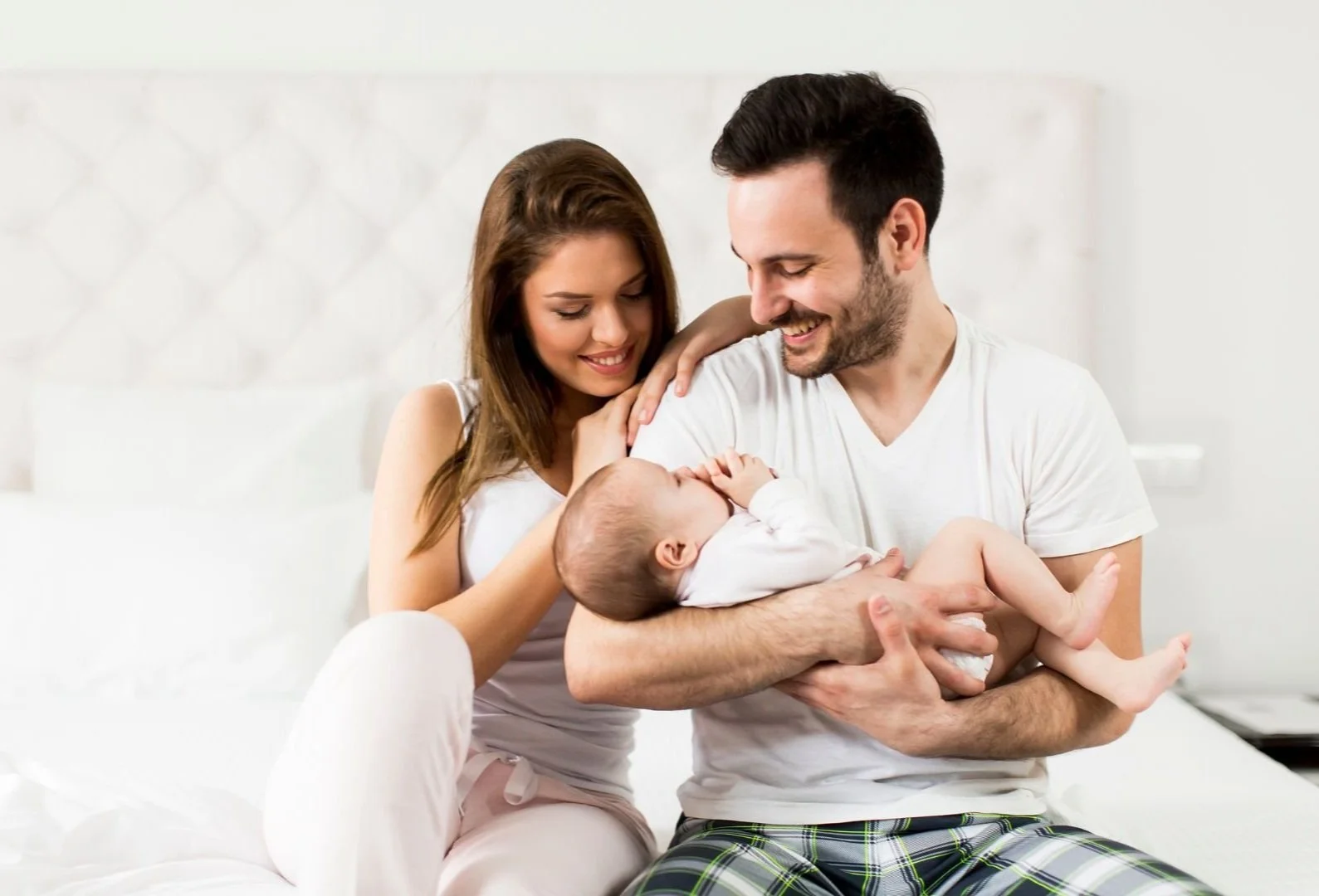 A happy couple sitting on a bed, smiling and looking at a baby in their arms.