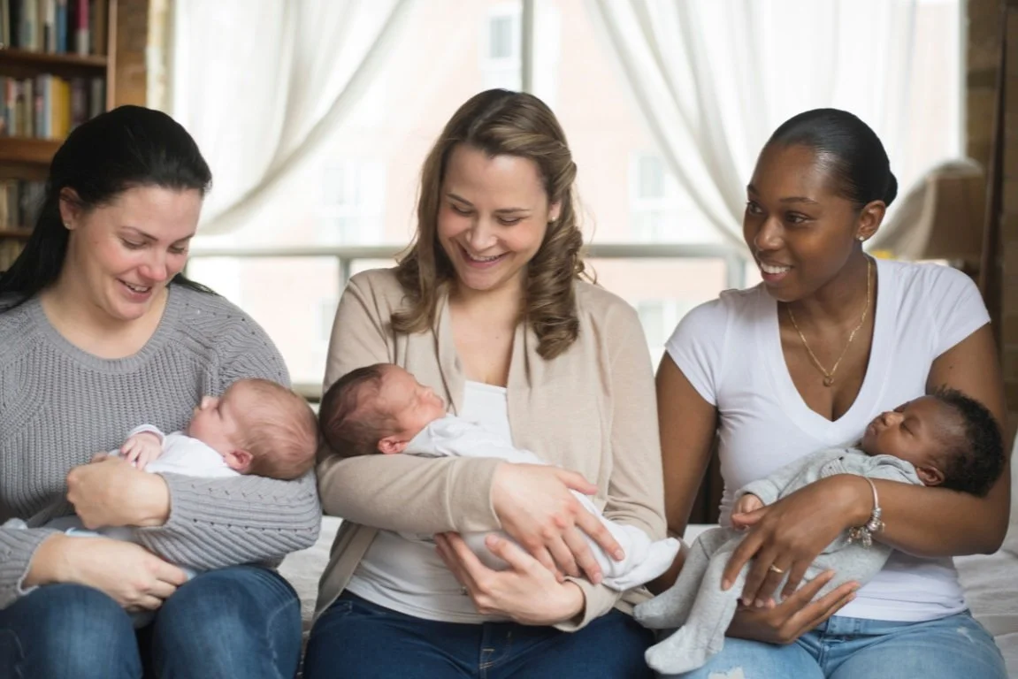 Three women sitting and holding infants, smiling and looking at the babies.
