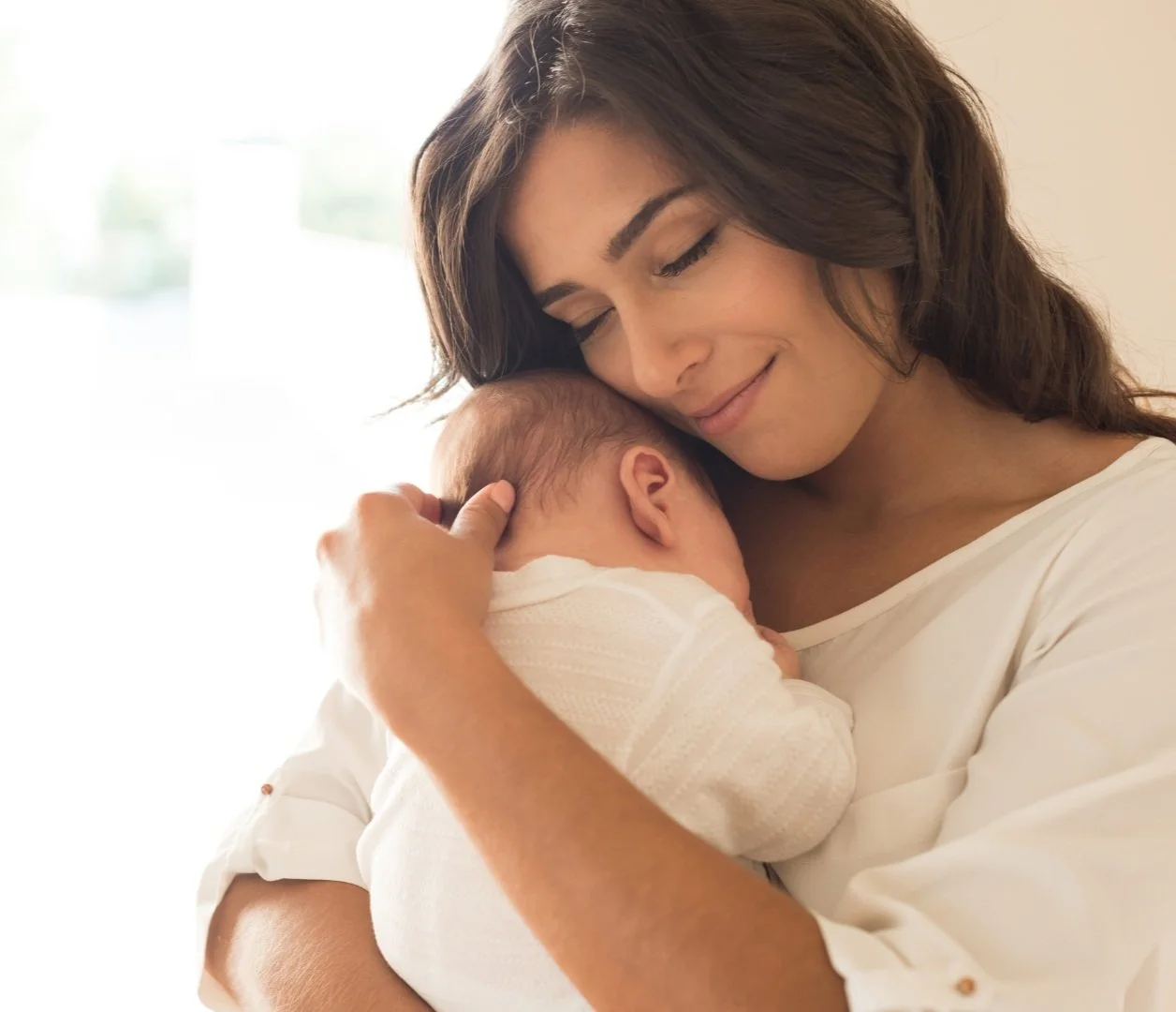 A woman holding a baby closely, both wearing white clothing.