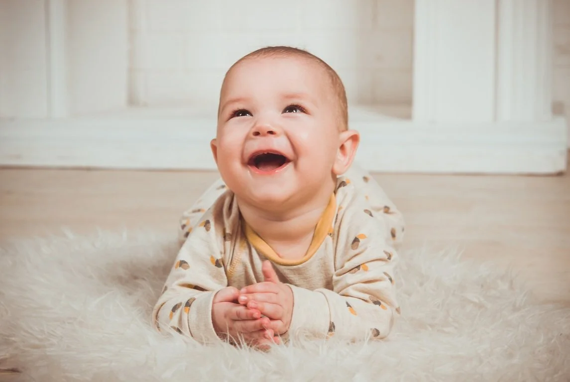 Happy baby lying on brown fluffy rug, smiling upwards, wearing light-colored onesie.