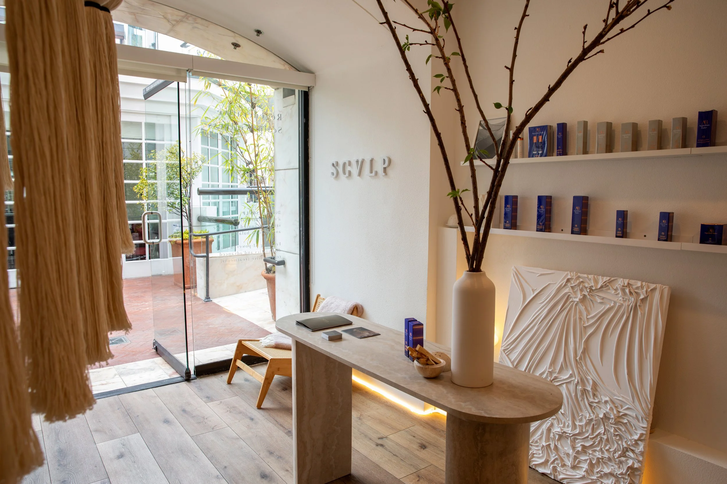 Interior of a modern spa or salon reception area with a marble reception desk, large white vase with branches, and a textured white art piece. Shelves with blue products are on the wall, and outside glass doors show potted plants and a patio area.