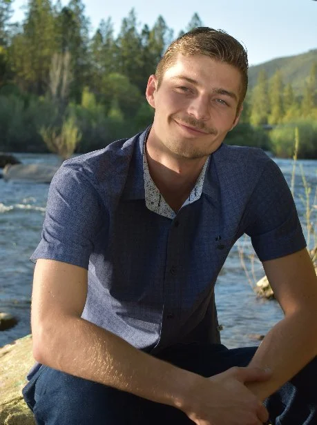 A young man with short hair and a mustache sitting outdoors by a river, smiling at the camera with a background of trees and hills.
