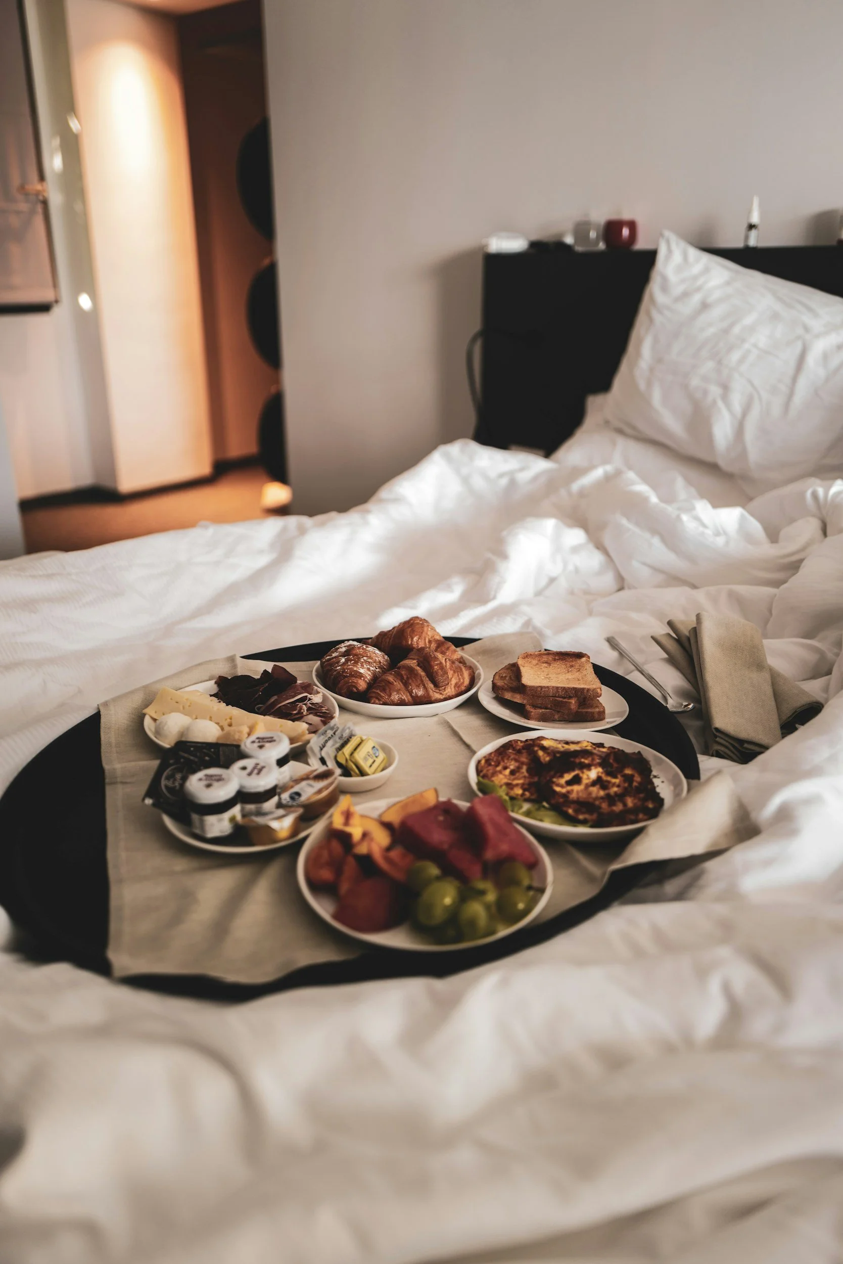 Breakfast in bed at a hotel or boutique hotel or motel or bed and breafast with croissants, toast, assorted cheeses, meats, small jam jars, fruit, and quiche on a tray on white bed linens.